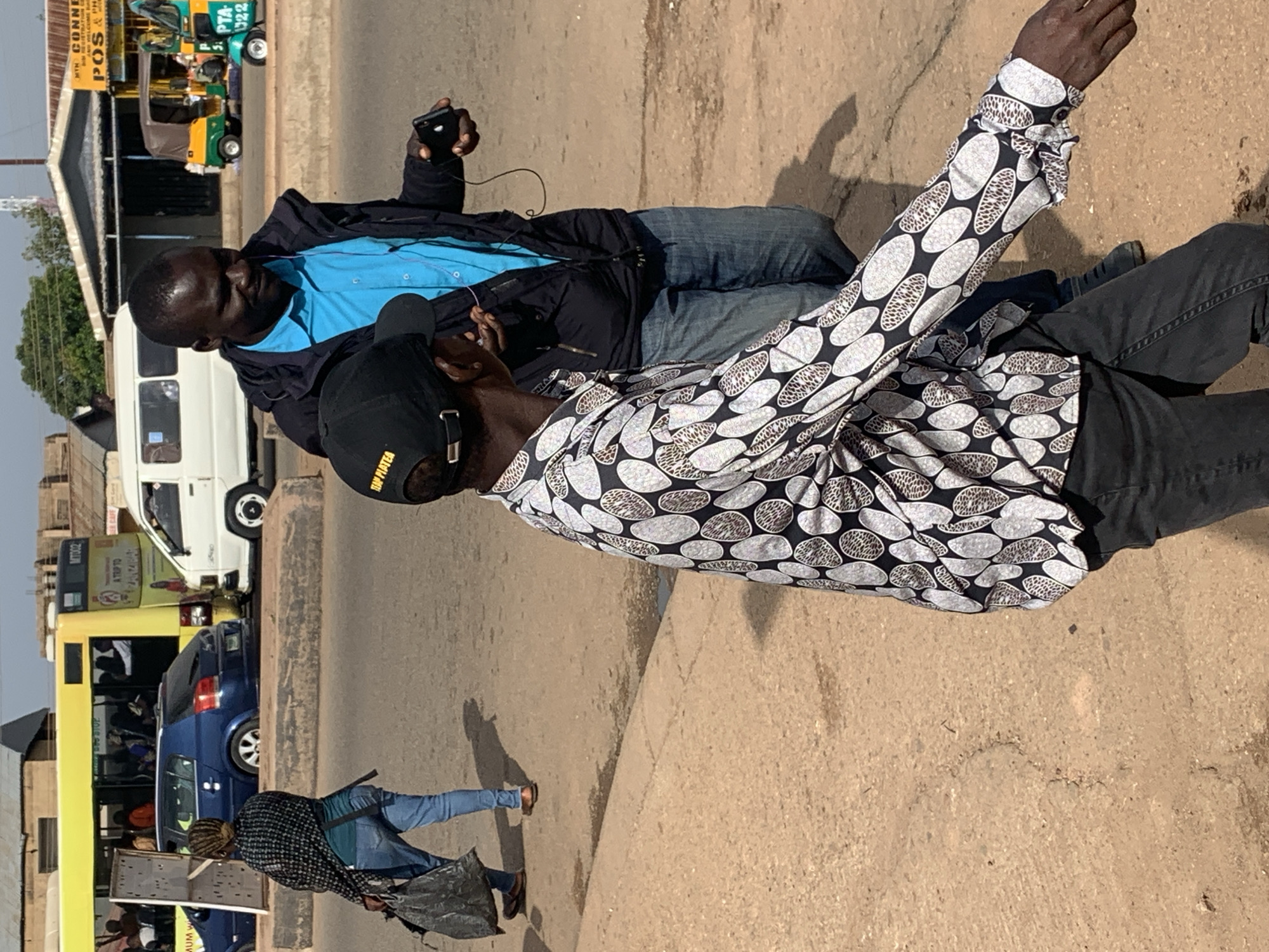 In Gura Top, Nigeria, two men are captured mid-stride on a dusty road. The man in the foreground, wearing a patterned shirt and a black cap, walks with his right arm extended. He is observed by another man in the background, who is dressed in a bright blue shirt and a dark jacket, holding a black device in his right hand. Behind them, various vehicles, including a yellow bus, a white van, and a three-wheeled motorcycle, are present, suggesting a bustling urban environment. The ground is unpaved and the sky is clear, indicating a sunny day.