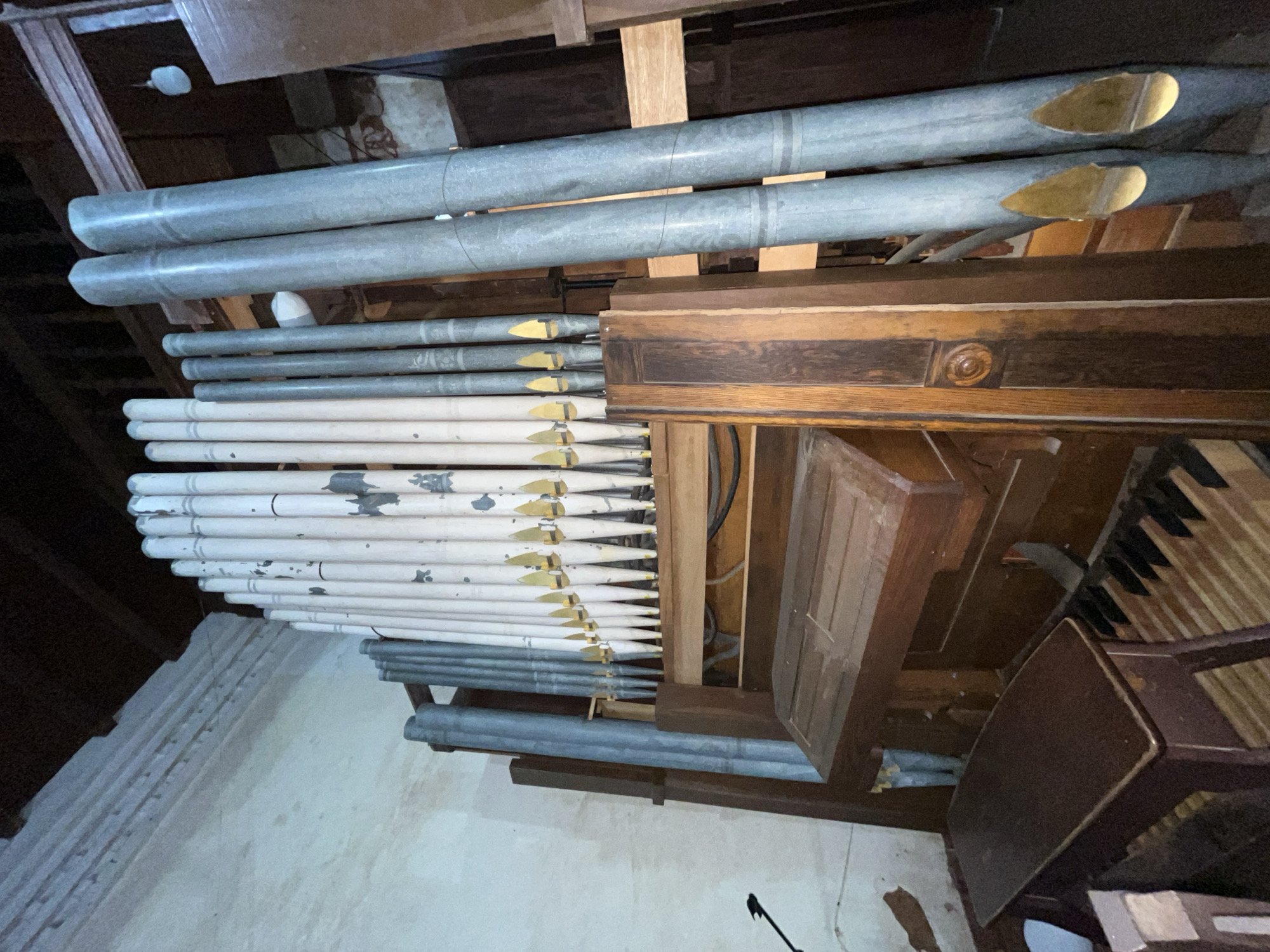 A close-up, tilted shot shows the internal workings of an organ, likely inside a building. A series of organ pipes, varying in size and color (some silver, some white with chipping paint), are arranged horizontally. The pipes have golden-colored mouths. To the right, there is a wooden console with visible keys, suggesting the location is a church or auditorium. The presence of wood and metallic organ pipes indicates a location in Unknown, Unknown.