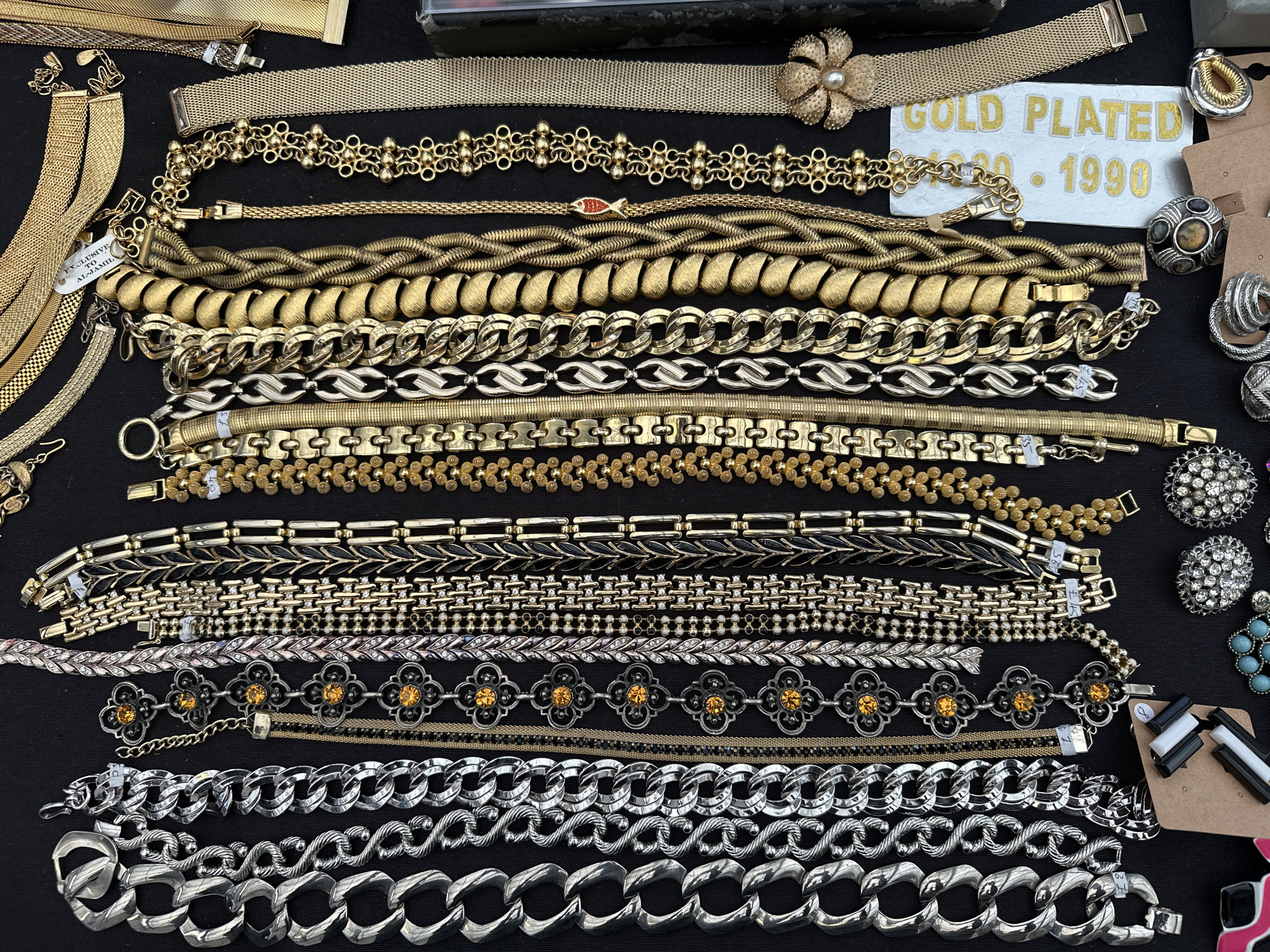The image displays a close-up of a collection of vintage costume jewelry, primarily bracelets, arranged on a black surface. The bracelets vary in material, color (gold, silver, black), and design, ranging from simple chains to more ornate pieces with embellishments like faux pearls and colored stones. A white sign with "GOLD PLATED - 1990" in gold lettering is visible. Some earrings are on the right side of the image. The setting appears to be a vendor's table at a flea market or antique fair.