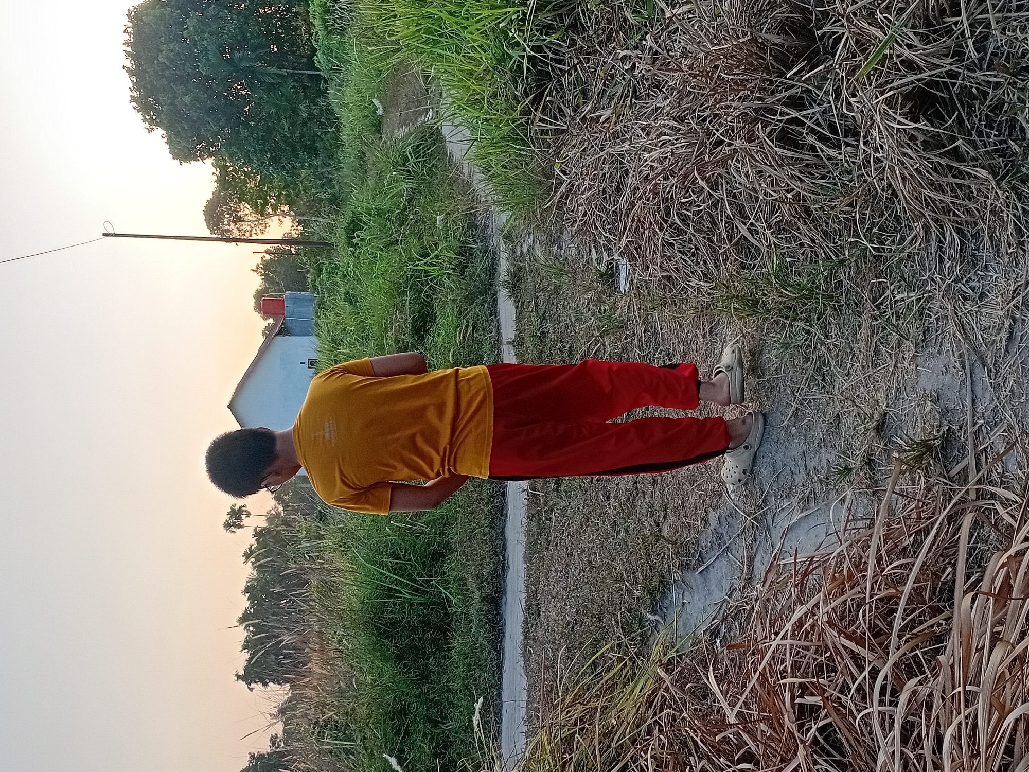 A young boy stands facing away from the camera in a grassy area, possibly a field or undeveloped lot, in Kasang Pudak, Indonesia. He wears a yellow t-shirt and red pants. Dry, brown grass and some green vegetation surround him. A narrow, dirt path winds through the scene. In the background, a white house with a red water tank is visible, along with trees and a utility pole against a pale sky, suggesting it's either early morning or late afternoon. The overall environment appears rural or semi-rural.