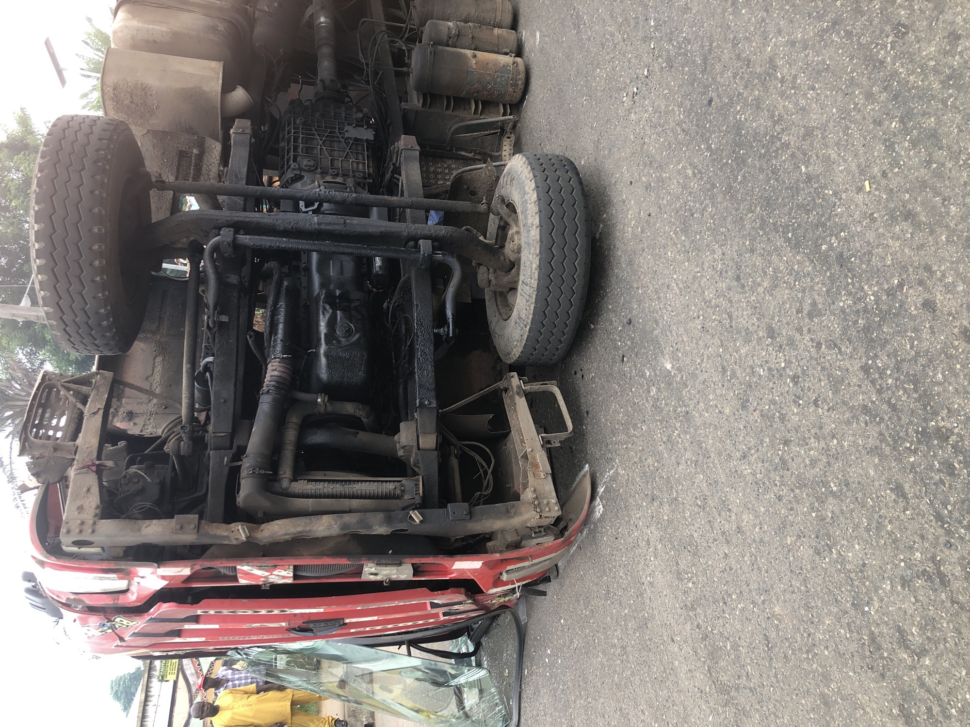 A red truck is overturned on a paved road, likely in Ikeja, Nigeria, with its tires facing upwards. The truck's undercarriage and engine are exposed, showing various pipes, gears, and tanks. A cracked windshield lies on the ground next to the cab. Two men in yellow and checkered shirts are standing on the left side of the road, observing the scene. The road appears to be asphalt, with some debris scattered around. The background shows foliage and buildings, suggesting an urban or suburban environment.