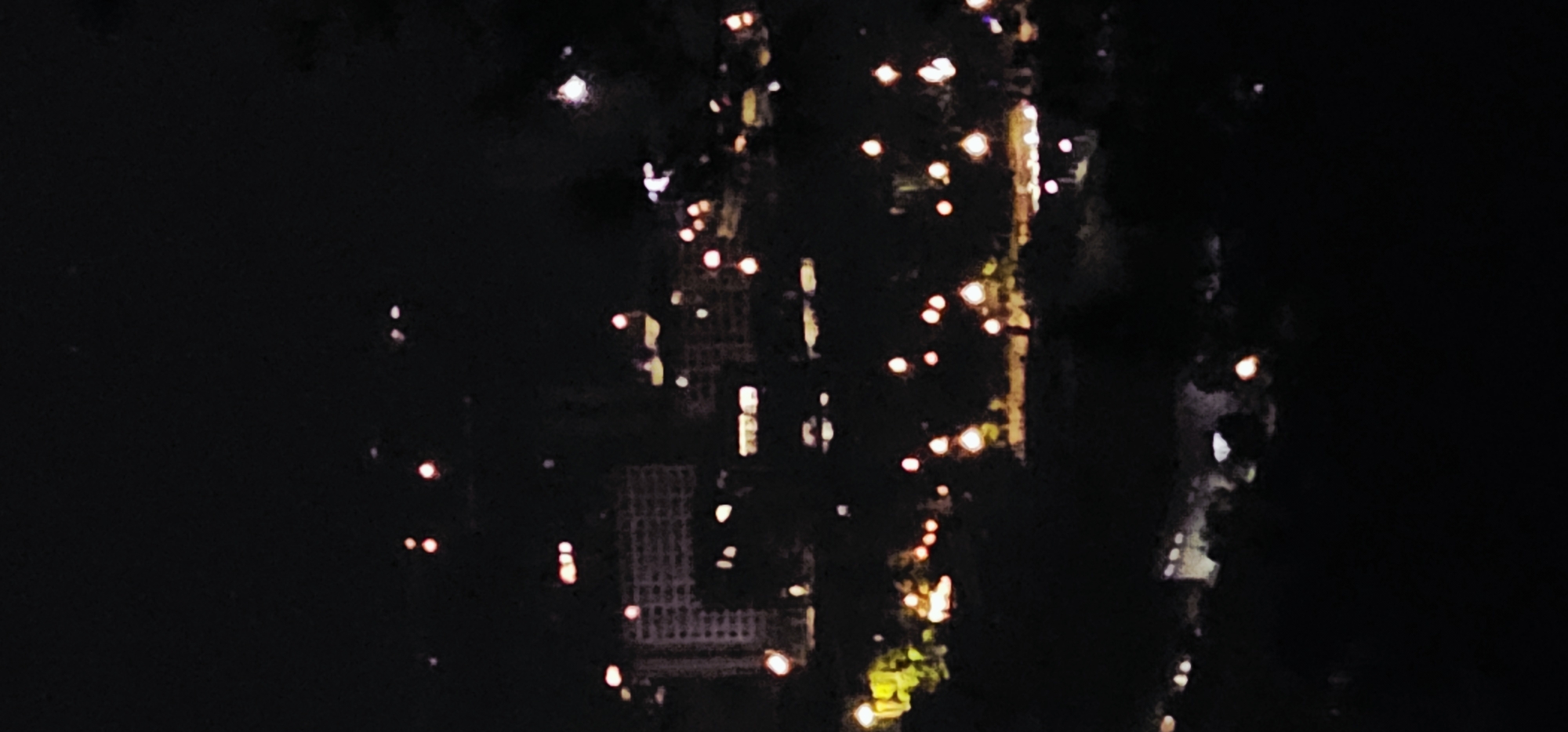 The image depicts a city skyline at night. The scene is dominated by darkness, with scattered points of light indicating buildings and artificial illumination. Two prominent structures resembling apartment buildings are visible in the middle ground, characterized by their rectangular shapes and multiple illuminated windows. The overall impression is of a residential or urban area at rest, with the lights suggesting activity within the buildings. The foreground is shrouded in darkness, obscuring any ground-level details. The background is also dark, with a few faint lights suggesting distant structures or the sky. The image is out of focus, giving the lights a soft, blurry appearance. The quality of the image suggests it may have been taken with a mobile device in low-light conditions. The specific location in Bishan, China, cannot be determined from the visual information provided.