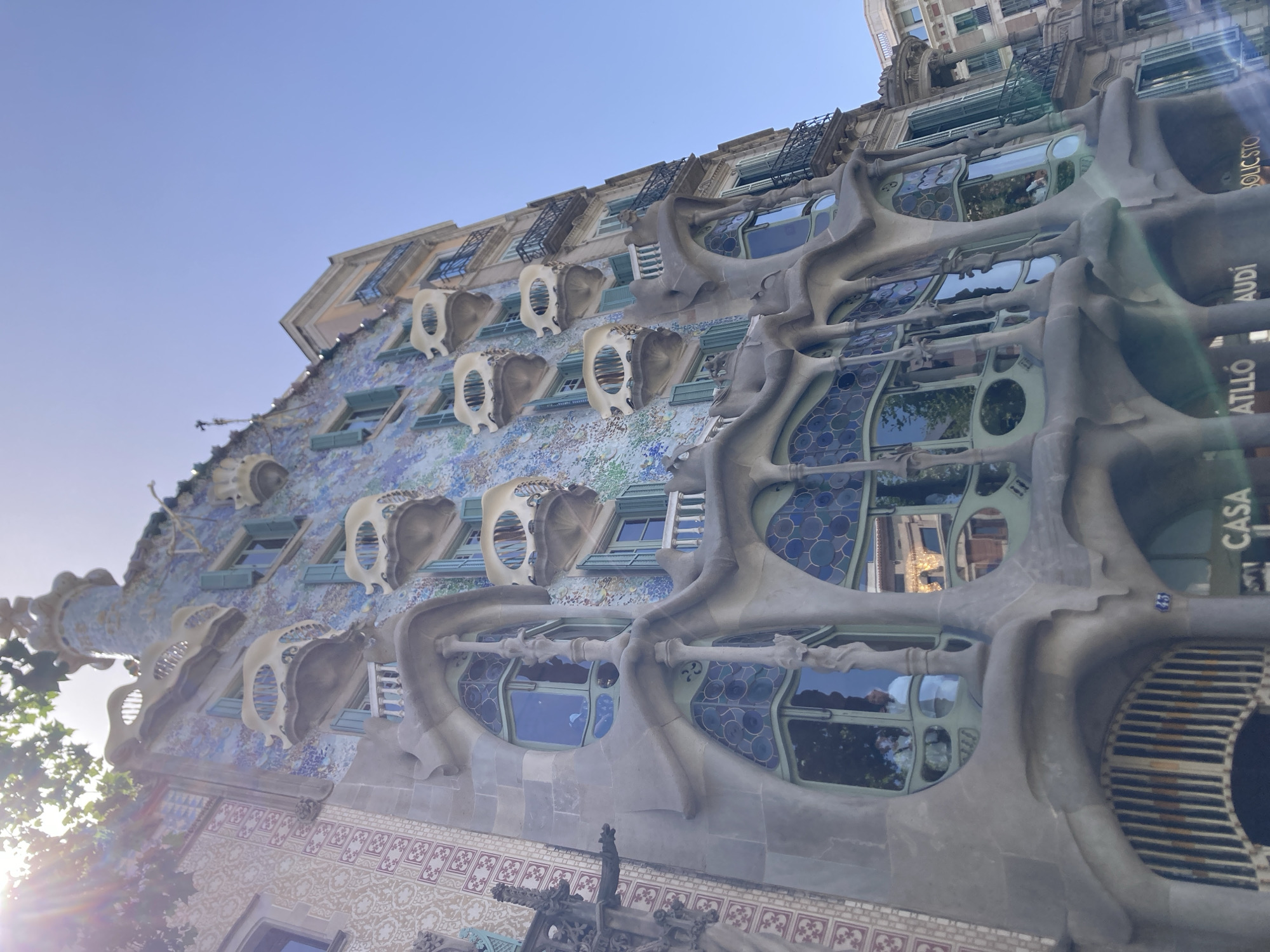 This media file captures a low-angle, wide shot of Casa Batlló, a renowned building in Barcelona, Spain, designed by Antoni Gaudí. The facade of the building is the primary subject, characterized by its organic, undulating forms, mosaic tiles in various shades of blue and purple, and distinctive bone-like balconies. Numerous windows are visible, some with intricate, multi-paneled glass, others framed by the building's characteristic sculptural elements. The architectural style is distinctly modernist and surreal, with flowing lines and an almost skeletal appearance to some of the external features.
Sunlight is glinting off the building, creating lens flare and highlighting the textures of the facade. The sky above is a clear, pale blue, suggesting it is daytime, likely morning or late afternoon due to the quality of the light and the soft shadows. Green foliage from trees is visible peeking from behind the building on the left, indicating the presence of surrounding greenery.
On the right side of the building, vertically oriented text is visible, including "CASA", "BATLLÓ", and "AUDI", indicating this is Casa Batlló and potentially a sponsor or related entity. The overall impression is one of awe at the unique and artistic architecture, set against a clear sky. No people are clearly visible in the frame, focusing the attention entirely on the building itself.