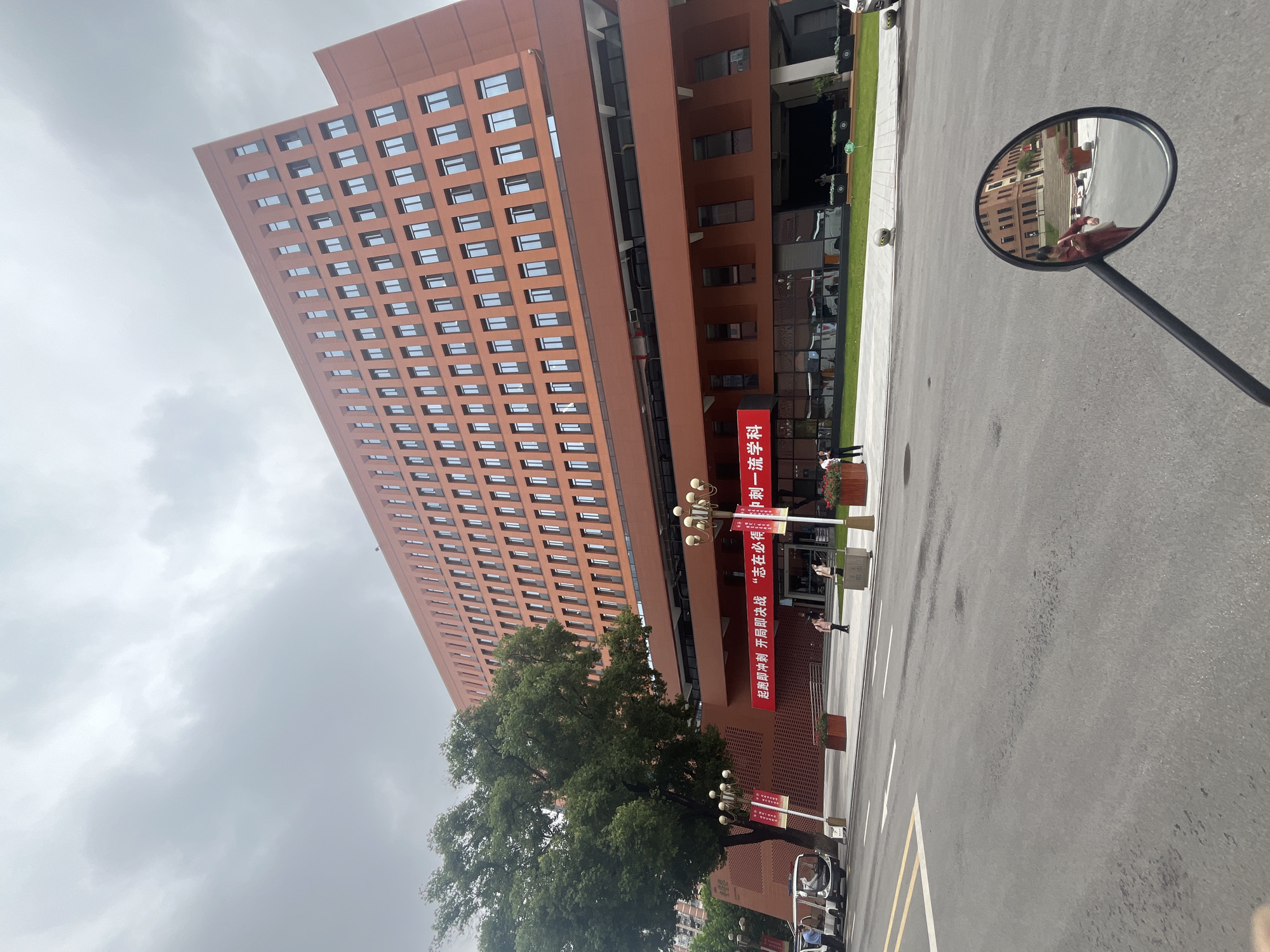 This is a wide-angle, eye-level shot of a modern, orange-brown brick building in Yuzhong District, China. The building has many windows, arranged in a regular grid pattern, and appears to be several stories high. In front of the building, there is a red banner with Chinese text. A large green tree stands to the left of the building, casting a shadow on the ground. A street is visible in the foreground, with a motorcycle's rearview mirror reflecting the scene, including a person wearing a red dress. There are also a few people walking on the sidewalk and a small white electric vehicle parked on the side of the road. The sky is overcast and gray.