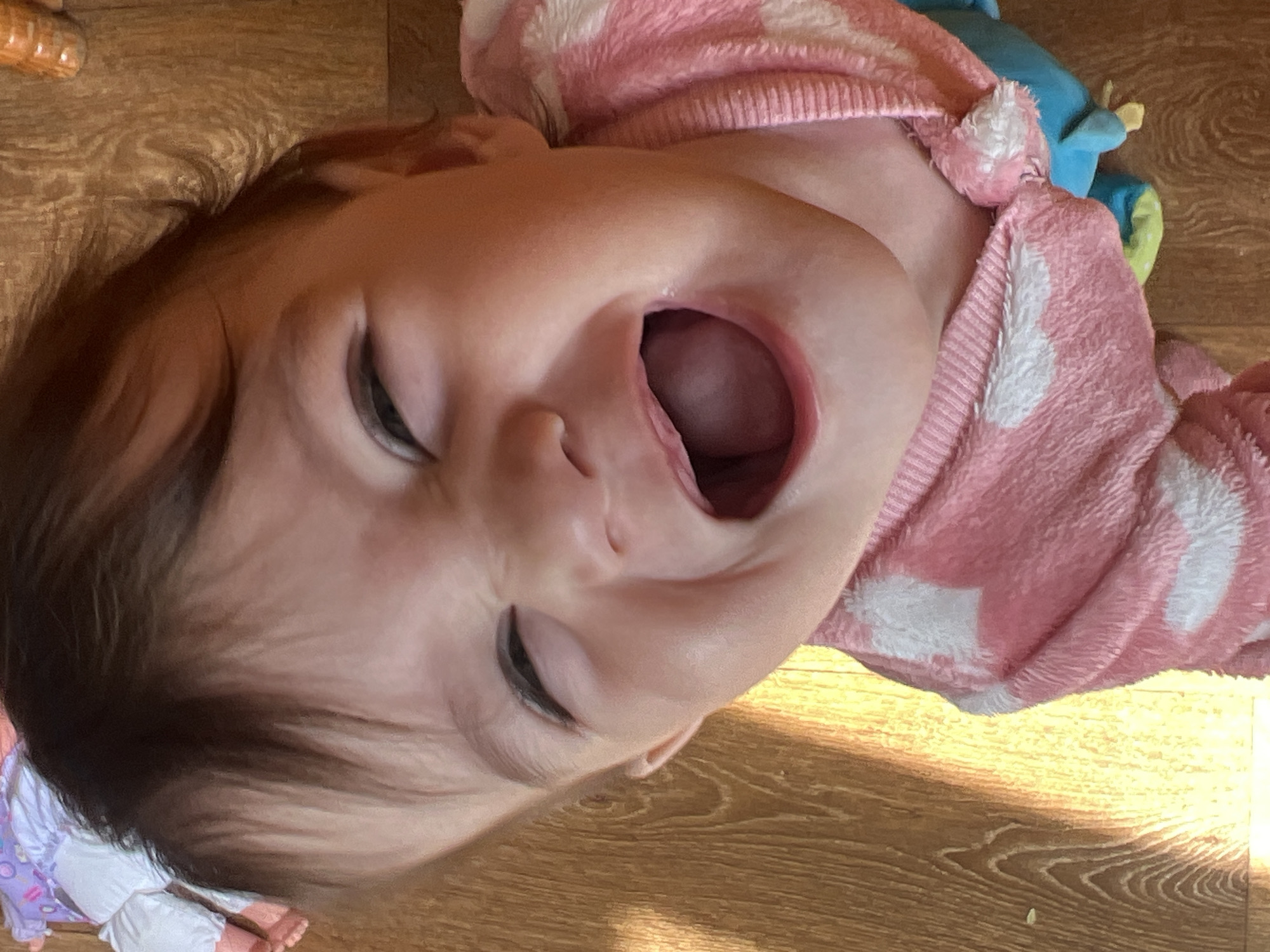 A baby with brown hair and fair skin is lying on their back on a wooden floor, wearing a pink and white patterned sweater. Their mouth is open, and their eyes are partially closed. A blue plush toy is near the baby. There are no contextual clues to indicate the location is in Elena, Bulgaria.