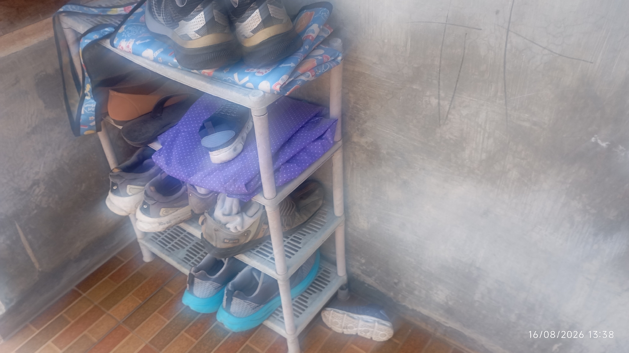 A three-tiered shoe rack is positioned against a concrete wall, with a tiled floor visible in the foreground. The shoe rack is filled with various footwear. On the top shelf, a pair of black and grey sneakers with white accents are visible. Below them, on the second shelf, a dark brown shoe and a blue and white sandal are placed next to a folded purple cloth with white polka dots. The third shelf holds two pairs of grey and white sneakers, one with a white sock protruding from it, and a pair of brown shoes. On the bottom shelf, a pair of blue and grey sneakers are displayed. A single blue and white shoe rests on the floor in front of the rack. The concrete wall behind the shoe rack has faint scratch marks. The image is dated 16/08/2026 at 13:38. This scene is likely located in Jatisobo, Indonesia, given the context.