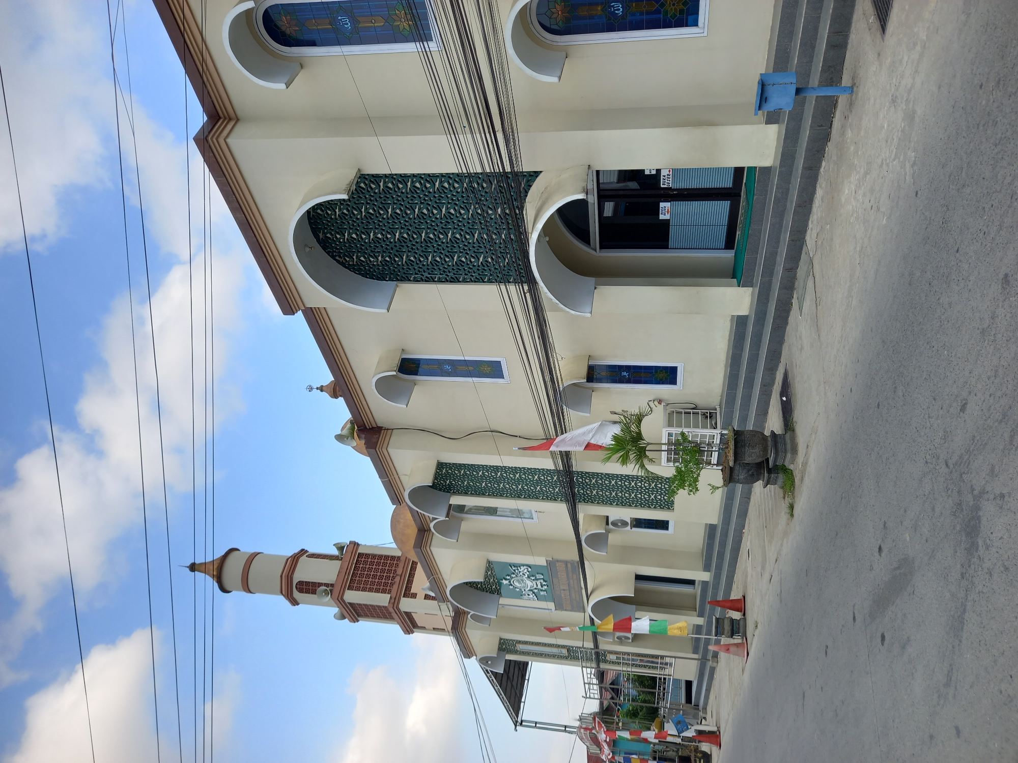 The image depicts the exterior of the Masjid Baitul Mukminin in Samarinda, Indonesia. The mosque is a light beige building with arched windows featuring blue stained glass with Arabic calligraphy. A tall, slender minaret with a copper-colored dome rises to the left of the main building. The mosque's facade also includes decorative green patterned screens above some of the arched entrances. In front of the mosque, there is a paved sidewalk with several potted plants and a few traffic cones on the road. A red and white Indonesian flag is prominently displayed near the entrance. The sky is partly cloudy, with power lines running across the upper portion of the image. The scene appears to be a daytime view of a street in a developed area, likely within the city.