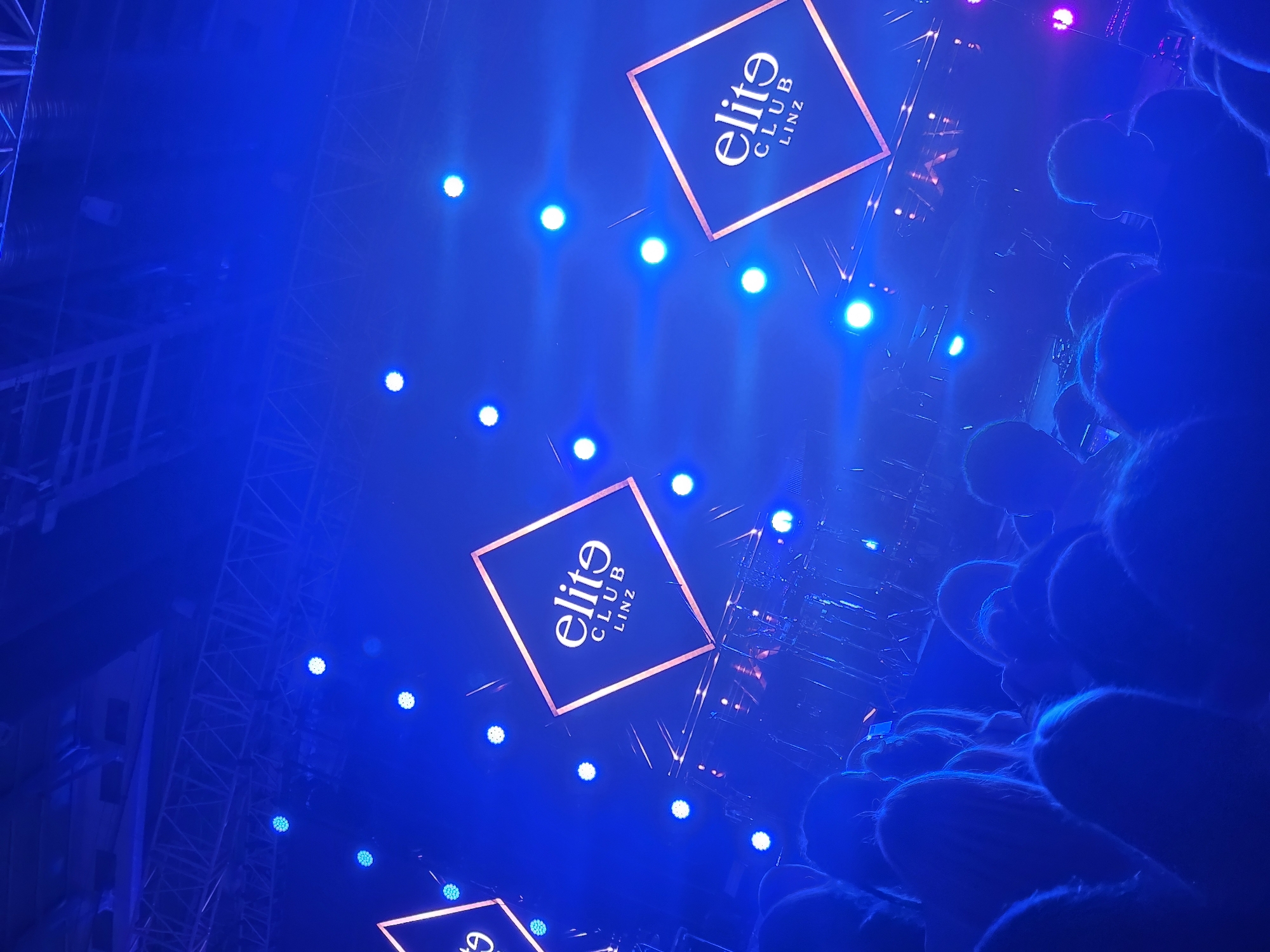 The image depicts a venue in Linz, Austria, identifiable by the "elite CLUB LINZ" signage. The scene is heavily illuminated with blue lighting, featuring multiple spotlights and the club's logo displayed in square frames. The setting appears to be a nightclub or entertainment venue, with stage rigging visible. The presence of a crowd is suggested by blurred figures in the foreground and background.