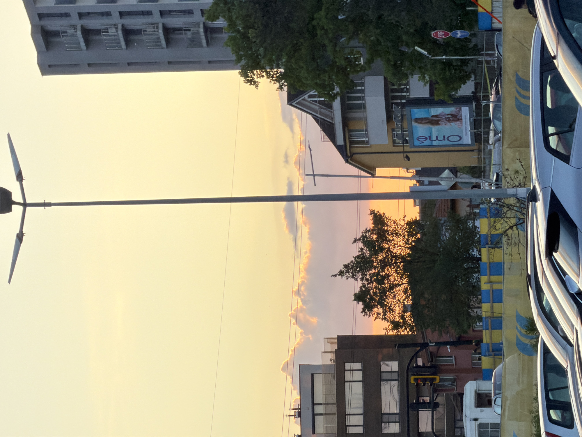 The image depicts a street scene in Sofia, Bulgaria during sunset. A tall street lamp stands on the left, its metal pole extending across the frame. The background shows buildings of various heights and architectural styles. A multi-story building with balconies is visible in the upper left. Another building features an "Omé" advertisement. A building is also showing with large windows. The sky is filled with orange and pink hues. Parked cars are on the right. Trees are scattered throughout the scene. Stop sign and other street signs are showing. A traffic light can be seen. The scene conveys a sense of urban life during the evening hours.