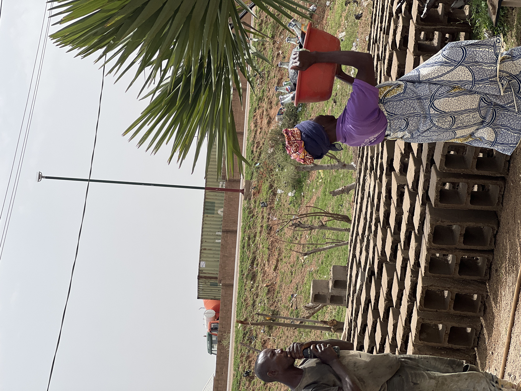 In Du, Nigeria, the image depicts two individuals in an outdoor environment. A woman wearing a blue headscarf, a purple t-shirt, and a blue and white patterned dress, carries a red bucket filled with glass bottles. The bucket is positioned above a stack of hollow concrete blocks. Nearby, a man wearing a dirt-stained shirt drinks from a bottle. In the background is a grassy area with sporadic brush and a building with shipping containers. A palm tree is present in the left side. Above, a utility pole and cables can be seen. The sky is overcast.