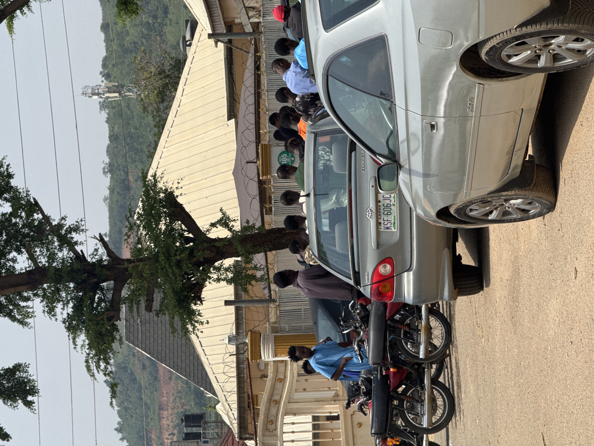 The image shows a street scene in Abuja, Nigeria, with a group of people gathered near several motorcycles and cars. The foreground features the rear of a silver Toyota Corolla and the side of another silver car. Behind these vehicles, a row of red and black motorcycles is parked. A young man in a light blue tunic stands near the motorcycles. Further back, a group of men are visible, some looking down, others interacting. A building with a light-colored, sloped roof and ornate metal gates is prominent in the mid-ground. A large tree with green foliage shades part of the scene. In the background, a telecommunications tower stands against a hazy sky, with a densely wooded hill behind it. The overall impression is of a busy urban environment, possibly a commercial or residential area.