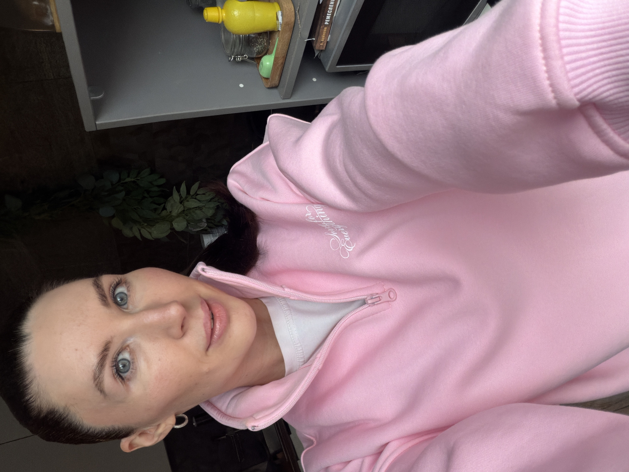 A fair-skinned woman with blue eyes and dark hair, wearing a light pink sweatshirt with a zipper and white lettering, is taking a selfie. Her face is turned to the left. Behind her are gray cabinets with items on top. There is also some greenery in the background. The image may have been taken indoors in a kitchen or similar room in Perm, Russia, although this is not explicitly stated.