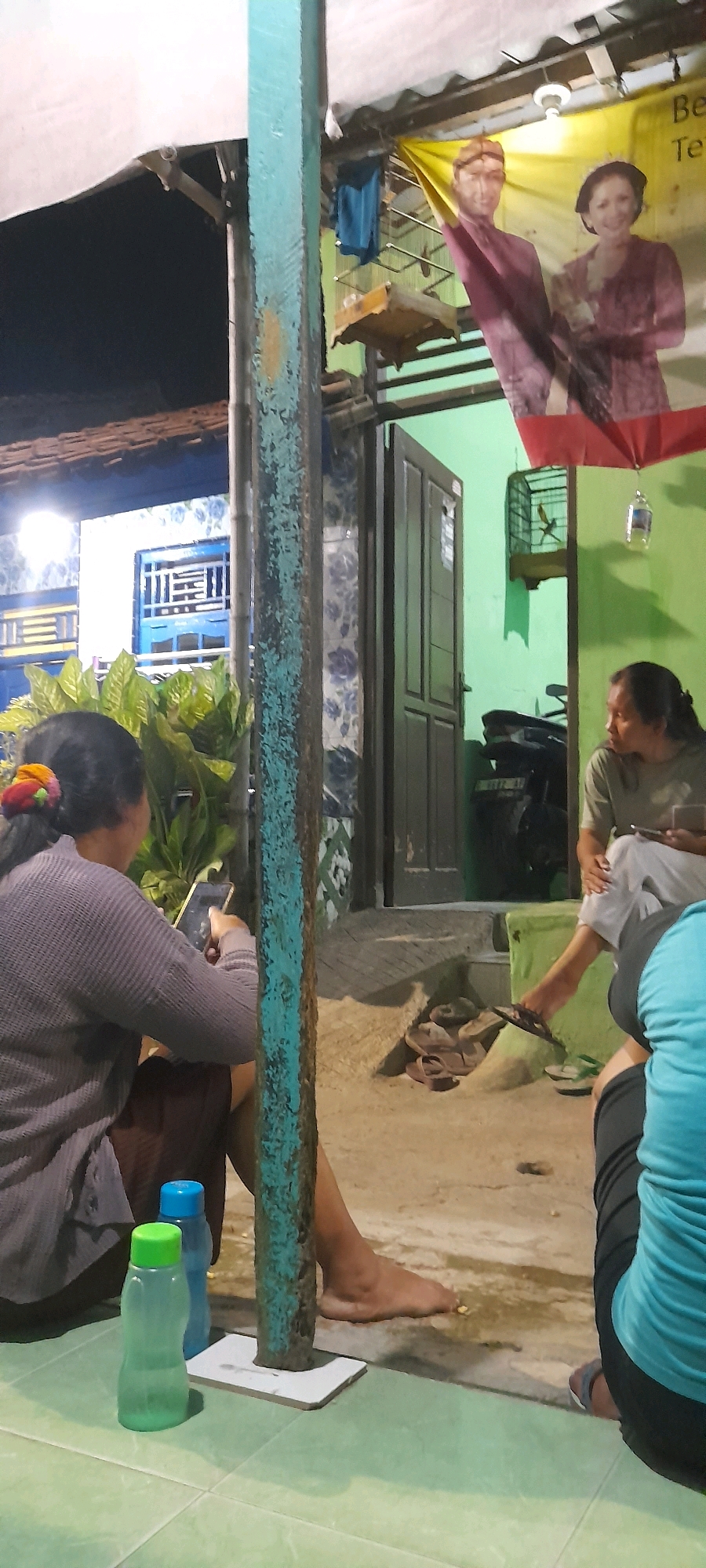 The image depicts an outdoor gathering in Tegal, Indonesia, likely in a residential or communal outdoor area. In the foreground, a person is seated and holding a smartphone, with two plastic water bottles nearby. To the right, another individual is seated, holding a smartphone. Behind them, a doorway is visible leading into a green-walled interior. A motorcycle is parked inside the doorway. Above the doorway, a banner features portraits of a man and a woman in traditional attire, possibly a wedding banner. A birdcage hangs from the ceiling. A turquoise-painted wooden pole is a prominent vertical element in the foreground, obscuring parts of the scene. The ground appears to be paved with tiles and concrete, with some scattered debris. The overall lighting suggests it is evening or late afternoon.