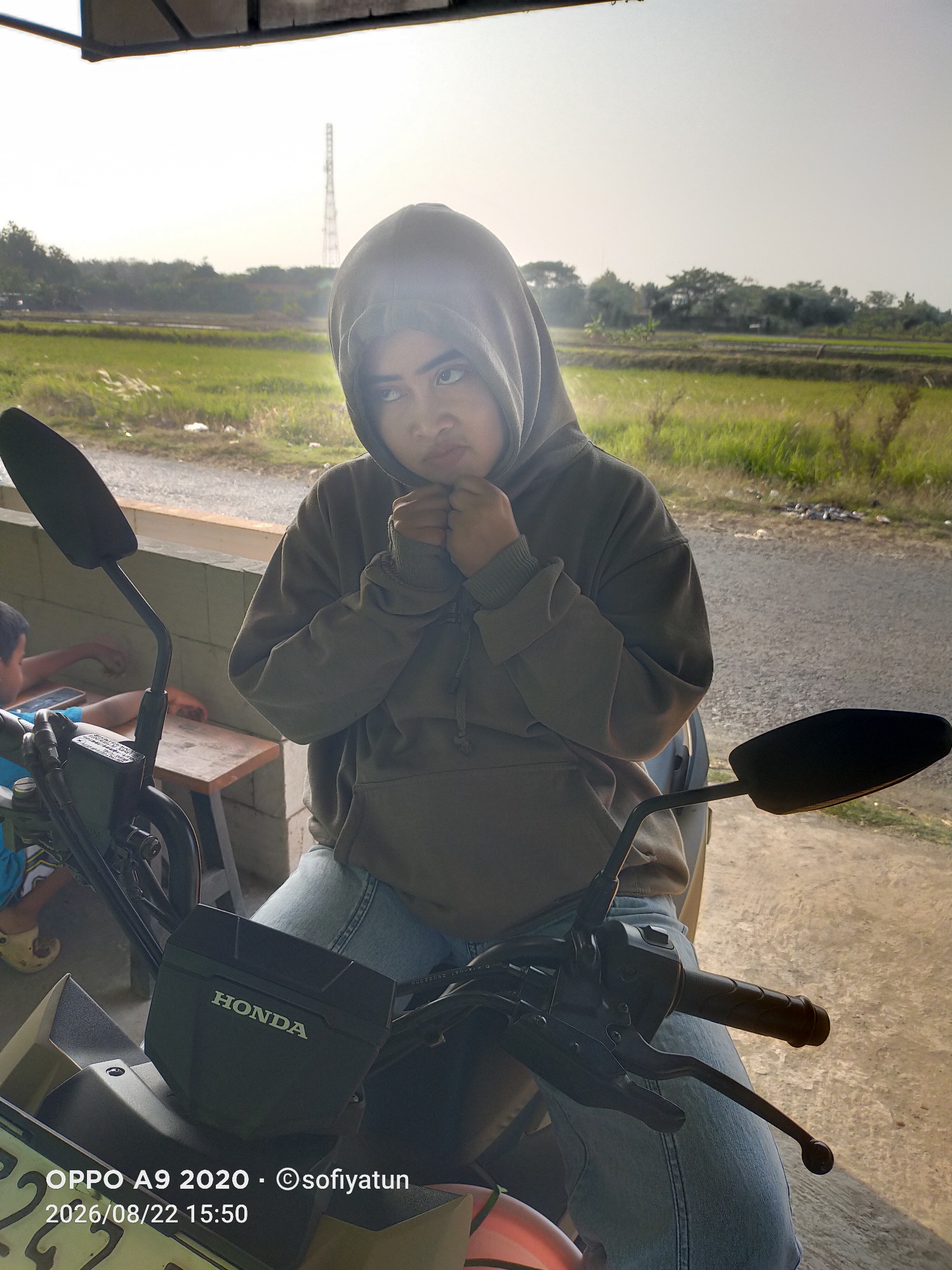 A young person wearing a grey hooded sweatshirt and blue jeans is sitting on a Honda motorcycle. Their hands are clasped in front of their chin, and they are looking up and to the left with a pensive expression. The motorcycle is parked on a paved surface next to a low concrete wall. Behind the wall, a field of green grass stretches into the distance, bordered by trees. A tall communication tower is visible in the far background. The sun is low in the sky, casting a warm glow over the scene. The timestamp "2026/08/22 15:50" and "OPPO A9 2020 - ©sofiyatun" are visible in the lower left corner of the image. The setting appears to be a rural area, consistent with the outskirts of Tayu, Indonesia.