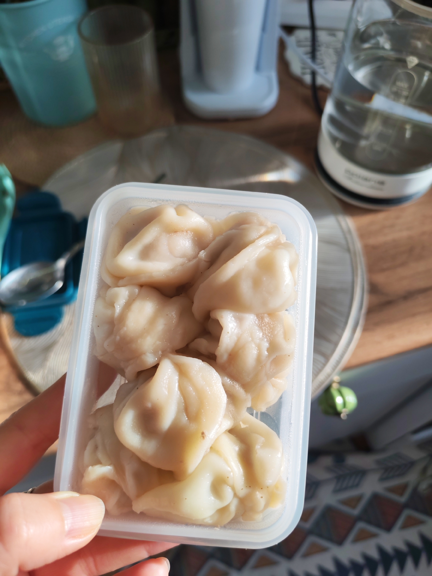 A person's hand is holding a clear plastic container filled with several cooked dumplings. The dumplings are pale, with visible folds and creases. The container is sitting on a table with a wooden surface. In the background, several objects are visible, including a teal cup, a clear glass, a white appliance, and a glass kettle filled with water. There is also a patterned fabric on the table. The scene appears to be indoors, in a domestic setting. The location is not specified as Izhevsk, Russia.