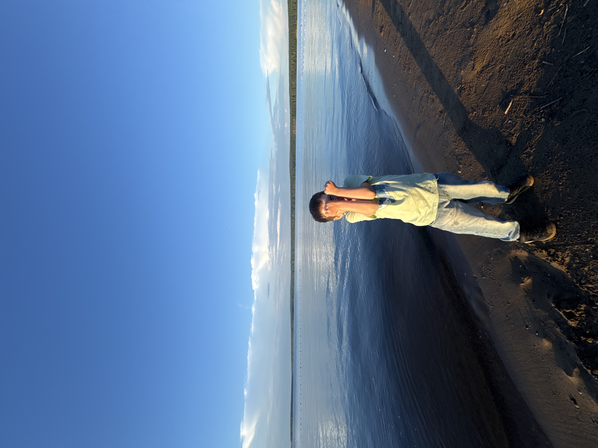 A young boy stands on a sandy beach, facing the viewer, with his arms raised and hands covering his eyes. He is wearing a light green t-shirt with a blue graphic and blue jeans. The beach is next to a large body of water, likely a reservoir or river, reflecting the bright blue sky and scattered white clouds. In the distance, a tree line forms the horizon. The overall lighting suggests it is late afternoon or early evening, with long shadows cast across the sand. The location is identified as the beach on the Kama River embankment in Chaikovsky, Russia.