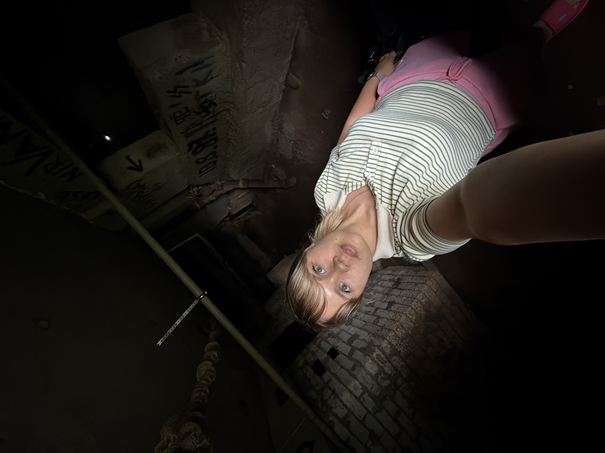 A woman in a striped shirt and pink pants stands inside an abandoned concrete structure. Markings, including "Nirvana" and other illegible graffiti, are visible on the concrete walls. A pipe runs along the wall, and the floor appears to be brick. The lighting is poor. The location is most likely the abandoned cooling tower of a defunct nuclear power plant in Chaikovsky, Russia.