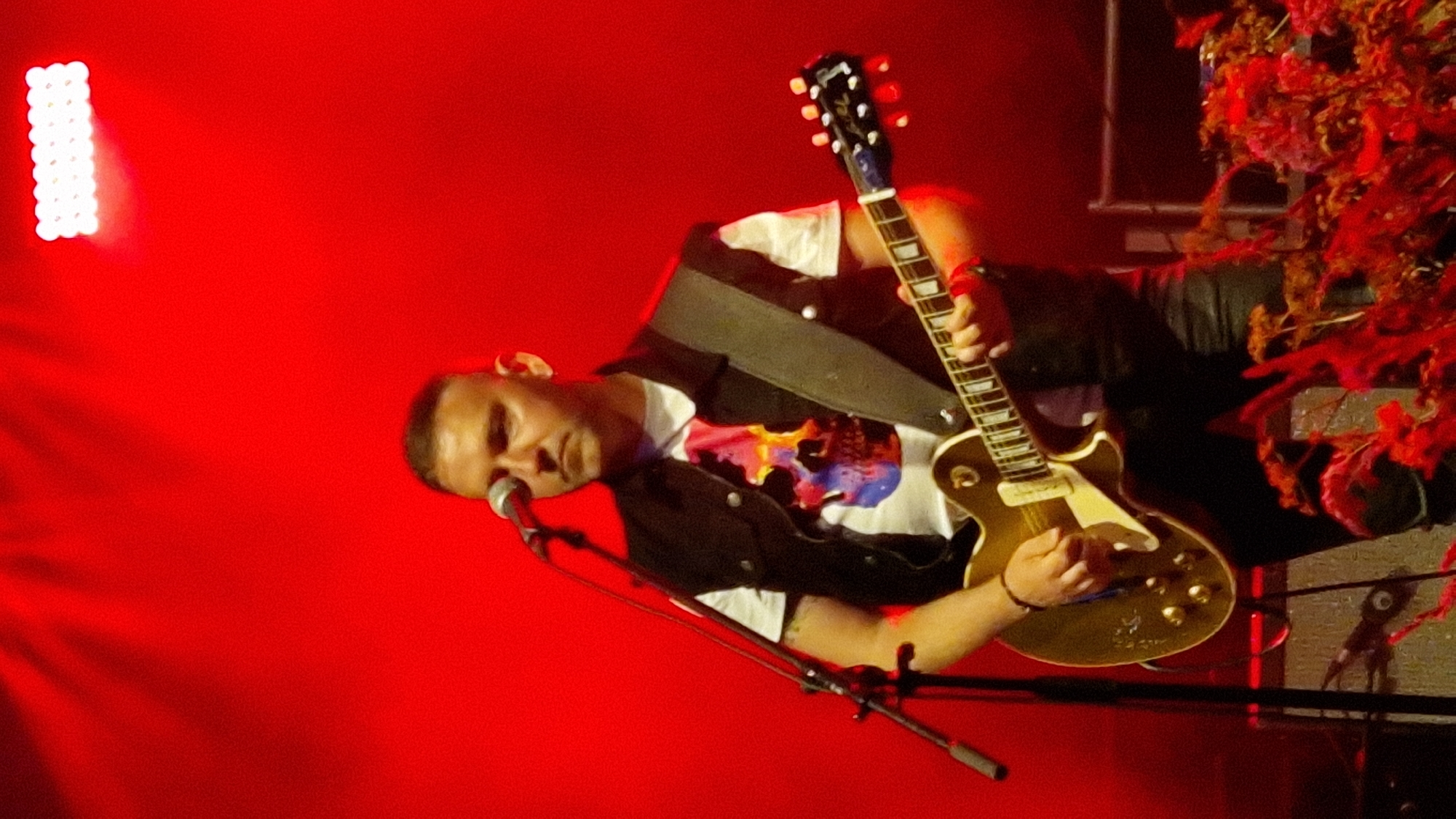 An image taken in Priekule, Latvia, depicts a man playing a golden electric guitar on a stage lit with red lighting. The man has a shaved head and is wearing a white t-shirt with a colorful skull graphic, a black vest, and dark pants. He is holding a microphone with his left hand and strumming the guitar with his right. The stage is equipped with a microphone stand, a guitar amplifier, and theatrical lighting. In the foreground, there are red flowers and foliage. The background is predominantly red, suggesting stage lighting or a colored backdrop.