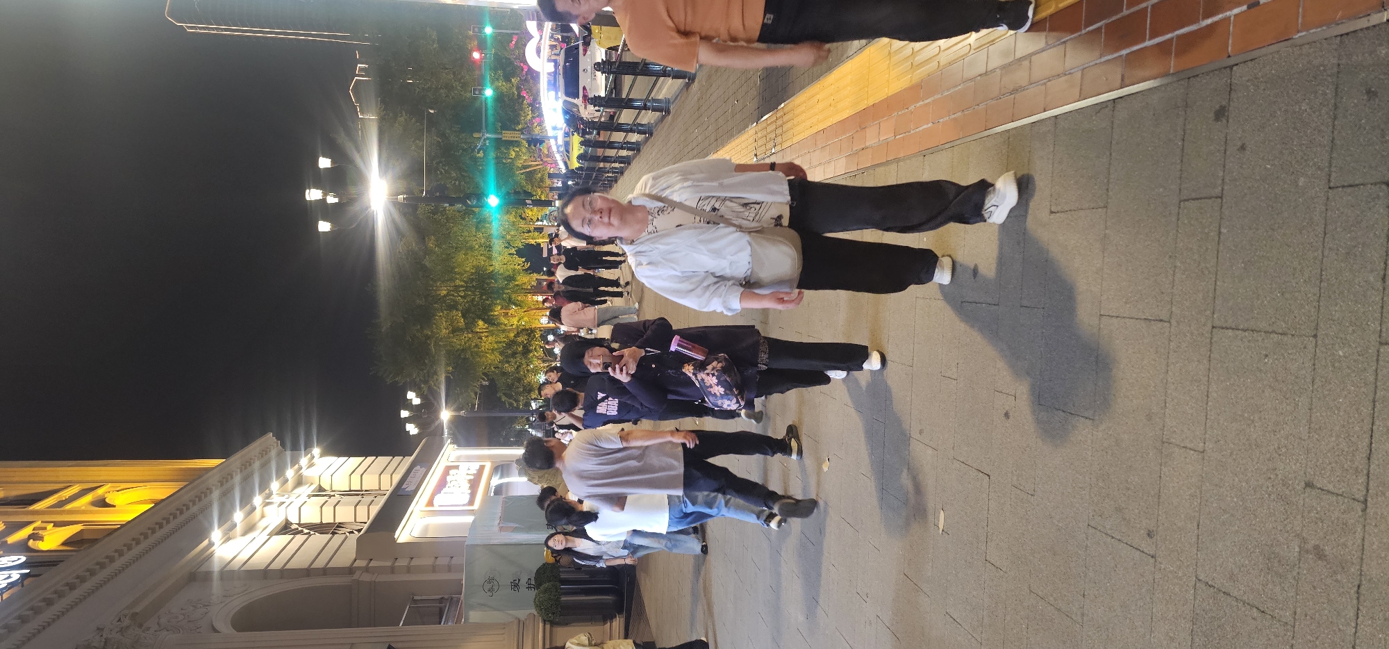 Captured at night in the Yuzhong District of China, the image shows a street scene with a mix of pedestrians. The architecture to the left includes a brightly lit, ornate building. Pedestrians walk along a paved area that transitions to brick. Street lamps illuminate the path, and greenery lines the route. The lighting suggests a festive atmosphere or decorative illumination, potentially indicating an event or special occasion. People are dressed casually, and some are engaged in conversation or looking at their phones.
