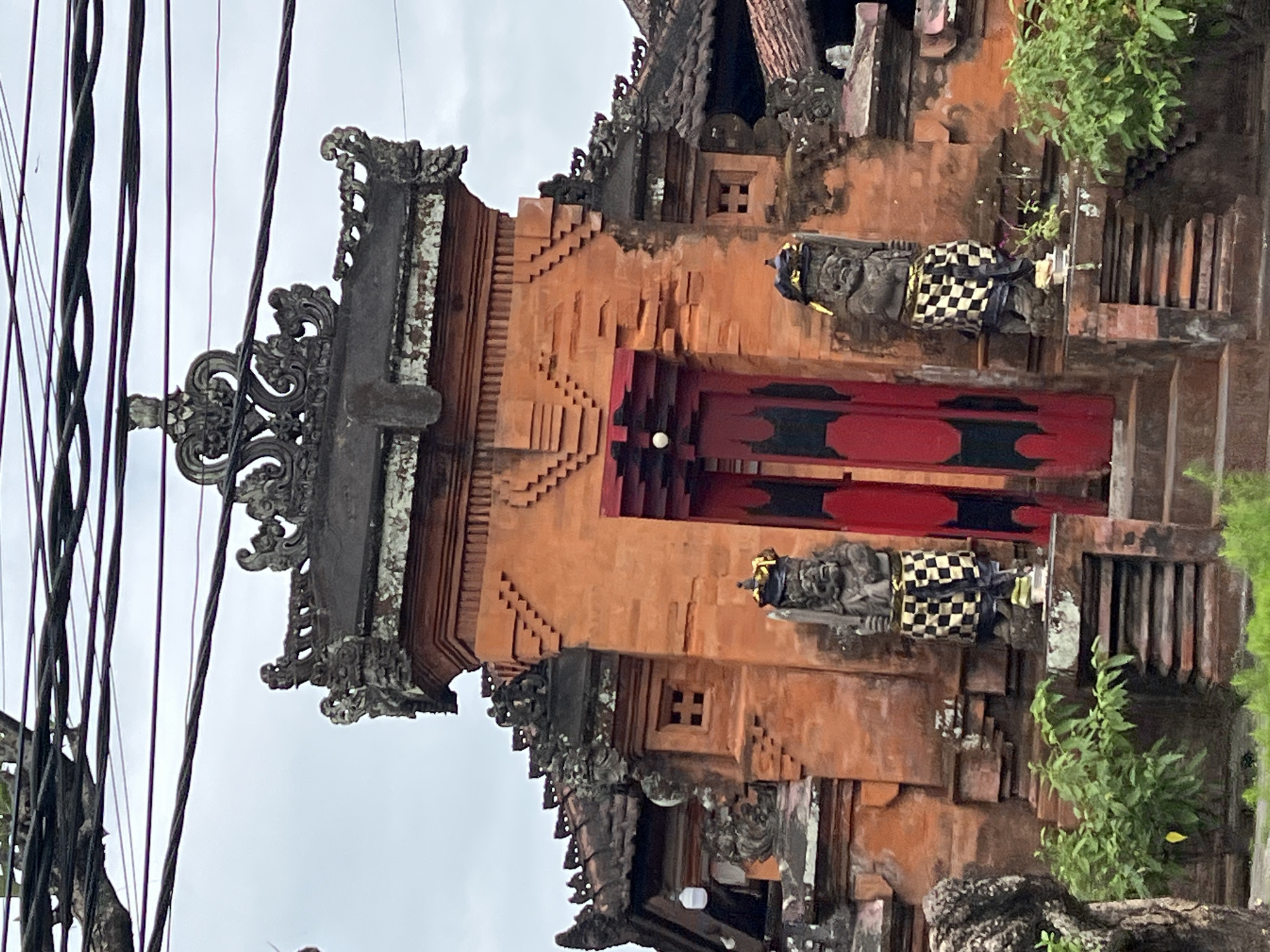 The image shows an ornate, traditional Balinese temple entrance in Denpasar, Indonesia. The temple is constructed primarily from reddish-orange brick, with intricately carved stone decorations adorning the rooflines and entryway. Two large stone statues, likely representing guardian figures, stand on either side of a central stairway leading to a bright red double door. These statues wear traditional checkered cloths around their waists and elaborate headdresses. The sky above is overcast, and a tangle of electrical wires and a tree branch are visible at the top of the frame, suggesting the temple is located in a developed area within the city. Lush green foliage grows around the base of the structure.