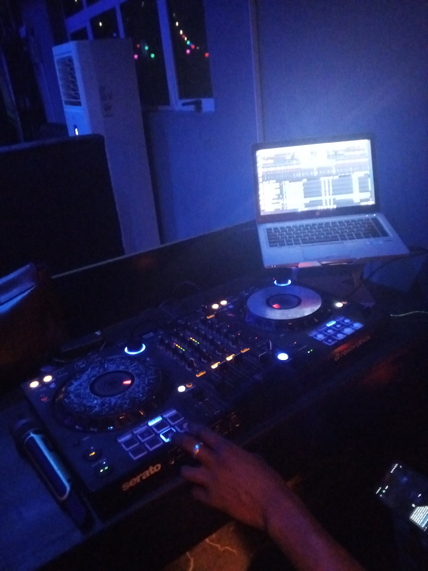 The image shows a DJ setup in a dimly lit environment, likely a club or a small venue. The DJ is interacting with a Serato DJ controller, with a laptop displaying DJ software positioned above the controller. A microphone is located to the left of the controller. A mobile phone lies to the right of the mixing deck. In the background, a portable air conditioning unit is visible near a window. The scene is bathed in blue light, contributing to the atmosphere. The image may have been taken in Lokoja, Nigeria.