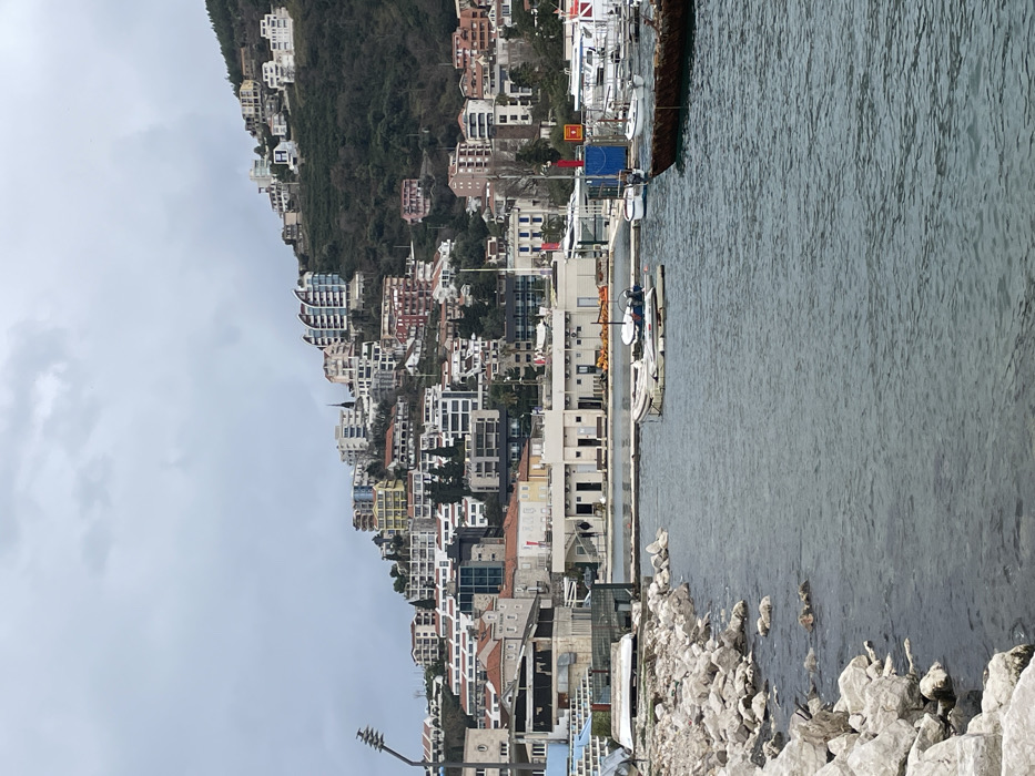 The image depicts a waterfront scene in Budva, Montenegro, under an overcast sky. In the immediate foreground on the left, a rocky embankment composed of large, light-colored stones extends along the water's edge. The water itself is calm, dark grey-blue, and shows minor surface ripples.
In the midground, a small white inflatable dinghy with an outboard motor is moored on a light-colored floating platform. To its right, a dark, heavily weathered or rusted boat is docked, and further right, several white boats, likely yachts, are moored within a marina marked by a red navigational structure and a yellow sign with a red border. On the adjacent shore, behind the floating dinghy, a series of light-colored, single-story buildings with flat roofs and dark entryways line the concrete path. Two indistinct figures are visible walking on this path. To the left of these buildings, a more industrial-looking structure with blue elements is visible, along with what appears to be an overturned white object or boat on land next to the rocky shore. A tall, metal light fixture or utility pole stands prominently on the far left.
The background is dominated by a steep, green hillside densely covered with a variety of buildings. These include traditional structures with red-tiled roofs and numerous modern multi-story residential or commercial buildings featuring diverse architectural styles, such as one prominent building with curved, wave-like blue and white balconies. A slender, dark spire rises above the buildings on the hillside, and a large white "H" sign is visible on a building higher up. A Montenegrin flag is also discernible in the marina area on the right.