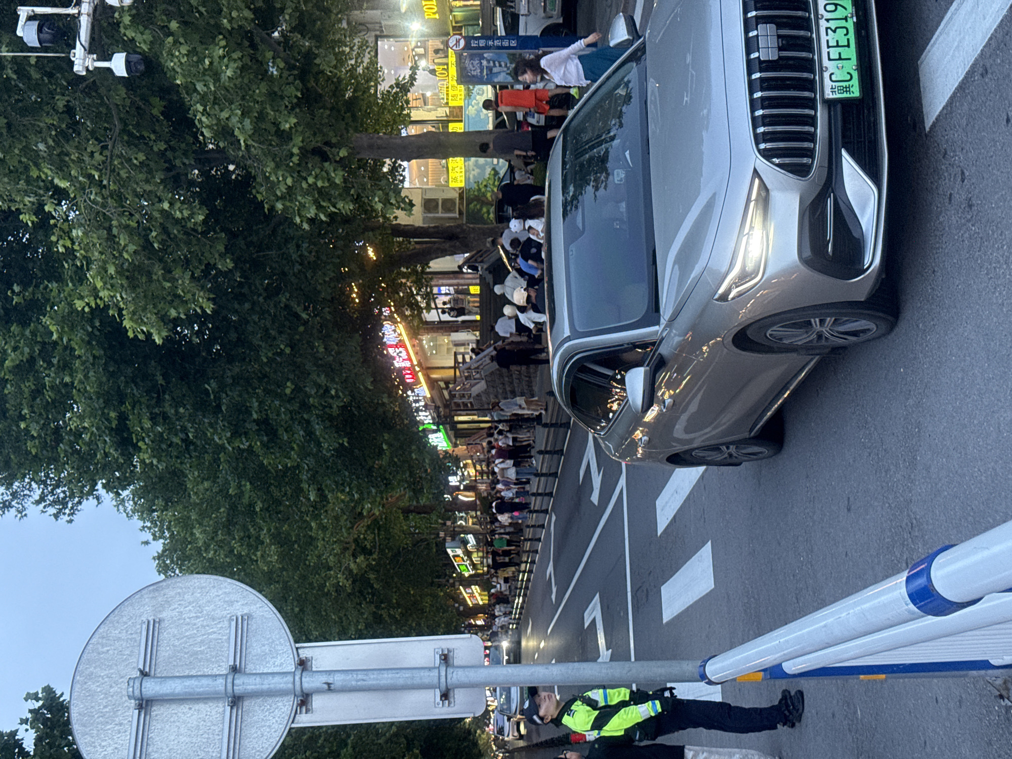 A silver sedan with license plate "冀C·FE3199" is stopped at a pedestrian crossing on a city street. A traffic police officer in a high-visibility yellow and green jacket stands to the left of the vehicle, facing forward. In the background, a crowd of people walks along a sidewalk lined with trees and illuminated by neon signs from shops and restaurants. The street appears to be in Qinhuangdao, China, based on the presence of Chinese characters on signs and the style of the license plate. Overhead, surveillance cameras are mounted on poles among the trees. The sky is overcast and the lighting suggests dusk or early evening.