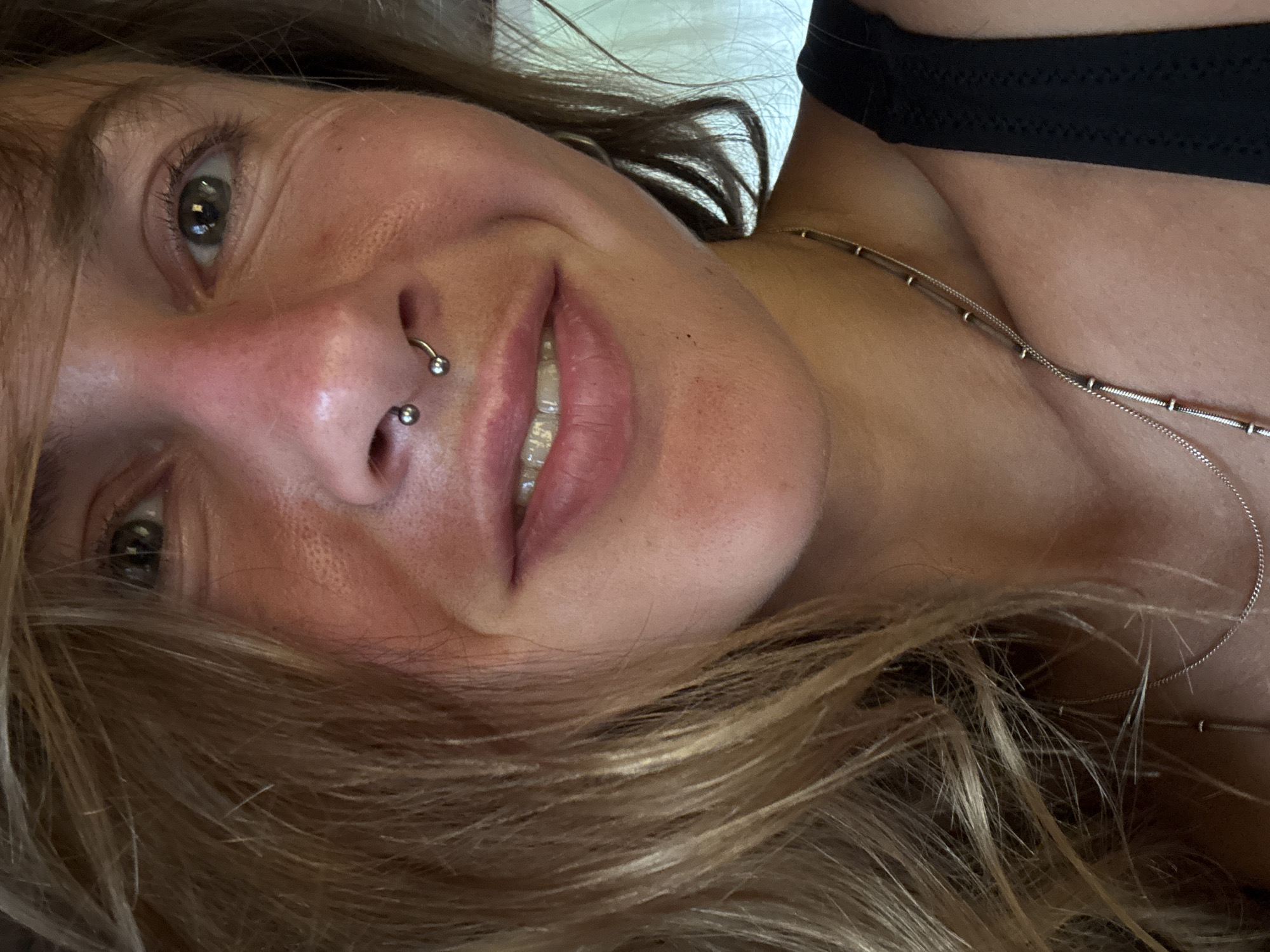 This is a close-up, vertically oriented, eye-level selfie of a young adult. The subject has fair skin with a smattering of freckles and is looking directly at the camera with a slight smile. Their hair, a mixture of blonde and light brown, frames their face. They have a septum piercing with two small silver balls and a circular barbell. The subject is wearing a black top and a delicate silver necklace. The background is a soft, out-of-focus white surface, suggesting an indoor setting. The lighting appears to be natural and soft, possibly indicating daytime. The overall mood is relaxed and intimate. No text is visible in the image.