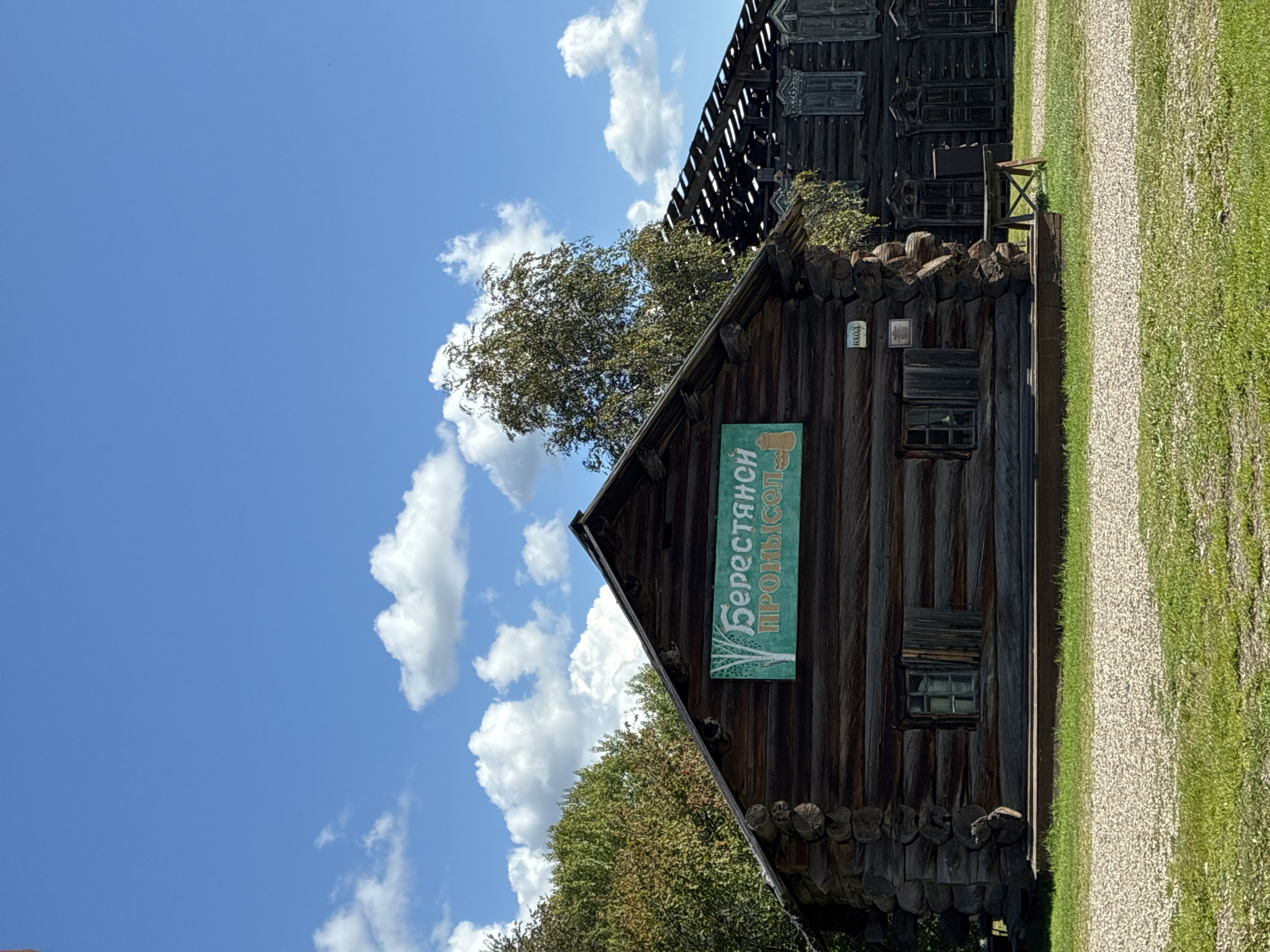 The image shows a wooden building with a sign that reads "БерестянО ПРОМЫСЕЛ" in Russian, which translates to "Birch Bark Crafts." The building appears to be part of an outdoor museum or historical village. To the right of this building, another wooden structure is visible, partially damaged or under construction, with its roof open to the sky. The foreground consists of a gravel path and green grass. The background features trees and a blue sky with white clouds. The scene is located in the Taltsy Museum, near Irkutsk, Russia, an architectural-ethnographic museum preserving historical Siberian wooden architecture.