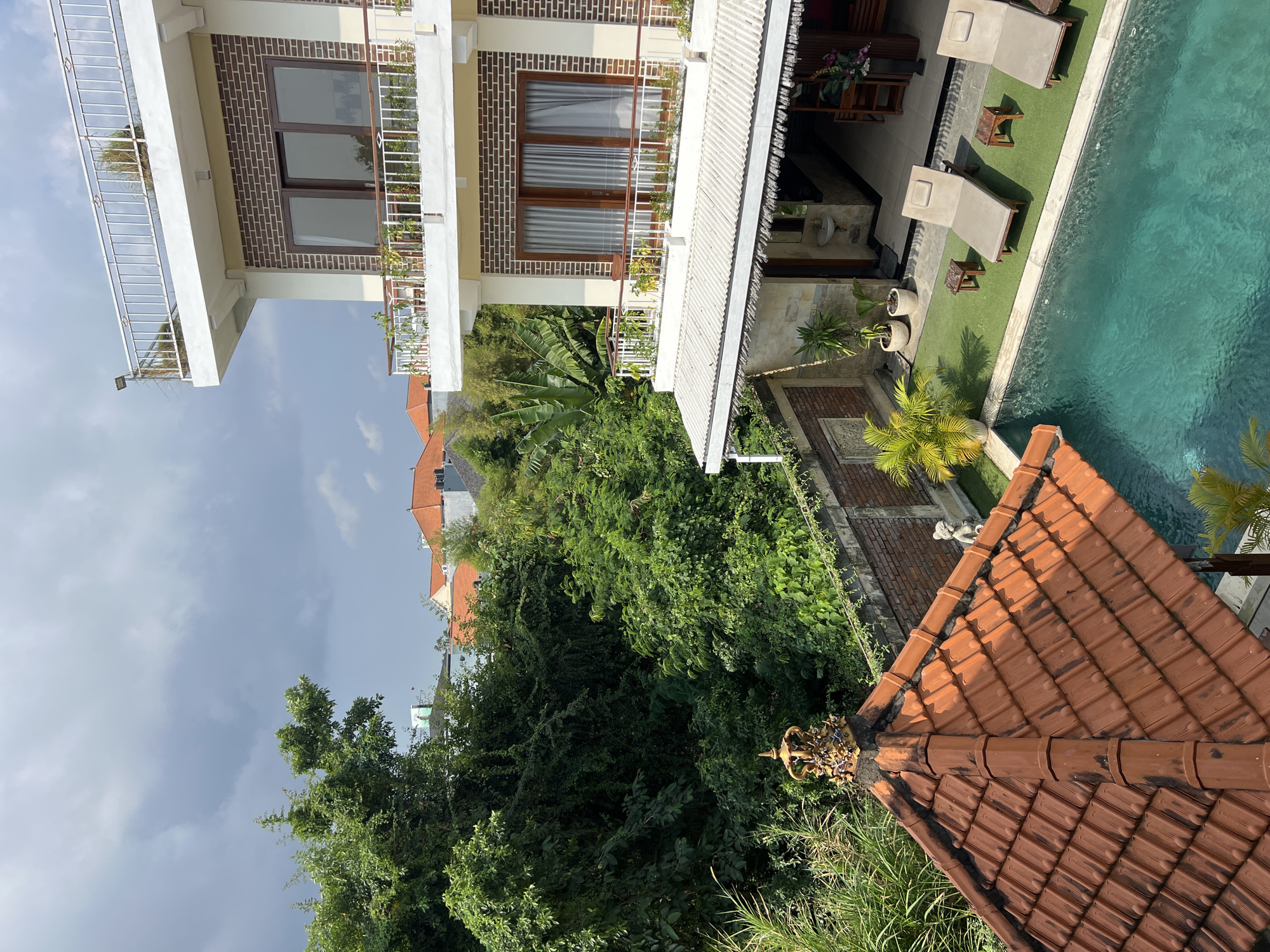 This high-angle, eye-level shot shows a resort in North Kuta, Indonesia. The resort features a multi-story building with white balconies and brown brick accents. The building overlooks a swimming pool surrounded by lush green foliage. A tiled roof structure with intricate golden crown-like embellishments is in the foreground. Two lounge chairs are positioned on the artificial grass next to the pool, with small wooden side tables. The scene is bathed in natural light under a cloudy sky, suggesting it is either morning or late afternoon. The overall ambiance is serene and tropical, typical of tourist destinations in Bali.