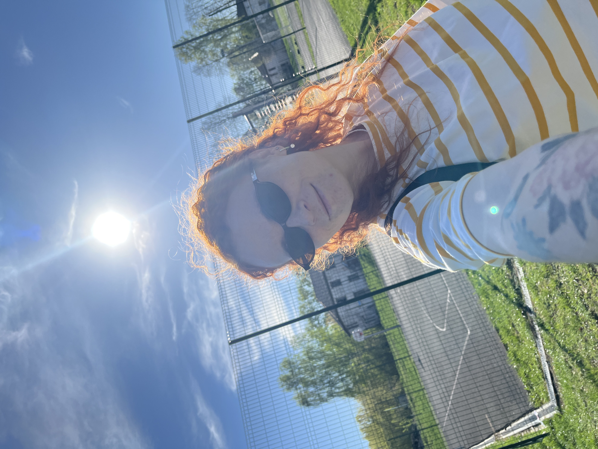 A low-angle outdoor shot depicts a person with red hair wearing sunglasses and a white and yellow striped shirt. The background shows a chain-link fence, green grass, trees, and a blue sky with some clouds and a bright sun. The location is possibly in the city of Chaikovsky, Russia.
