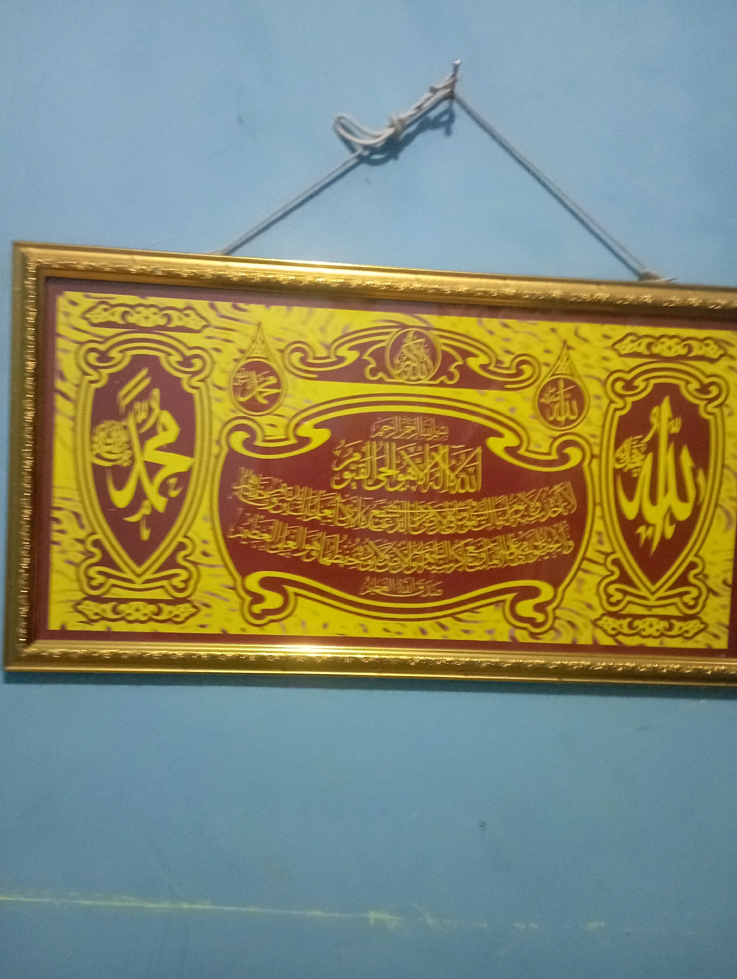 The image shows a framed Islamic calligraphy artwork hanging on a light blue wall. The artwork is rectangular and framed in gold. It features a yellow background with intricate red and gold Arabic script and motifs. On the left side, there is a shield-like design with yellow calligraphy on a red background, which appears to be the name "Muhammad". On the right side, there is a similar shield-like design with yellow calligraphy on a red background, likely representing the word "Allah". In the center, there is a larger section of red calligraphy on a yellow background, surrounded by decorative red and yellow swirls and shapes. The artwork is suspended by a thin rope tied to a fixture on the wall, creating a triangular suspension. The wall is plain and appears to be painted a matte, light blue color. The lighting is somewhat dim, casting a slight shadow from the rope. The scene is a close-up of the artwork and the wall it hangs on, with no other elements of the environment visible. This artwork is likely located in a private residence in Senangsari, Indonesia, given the context.