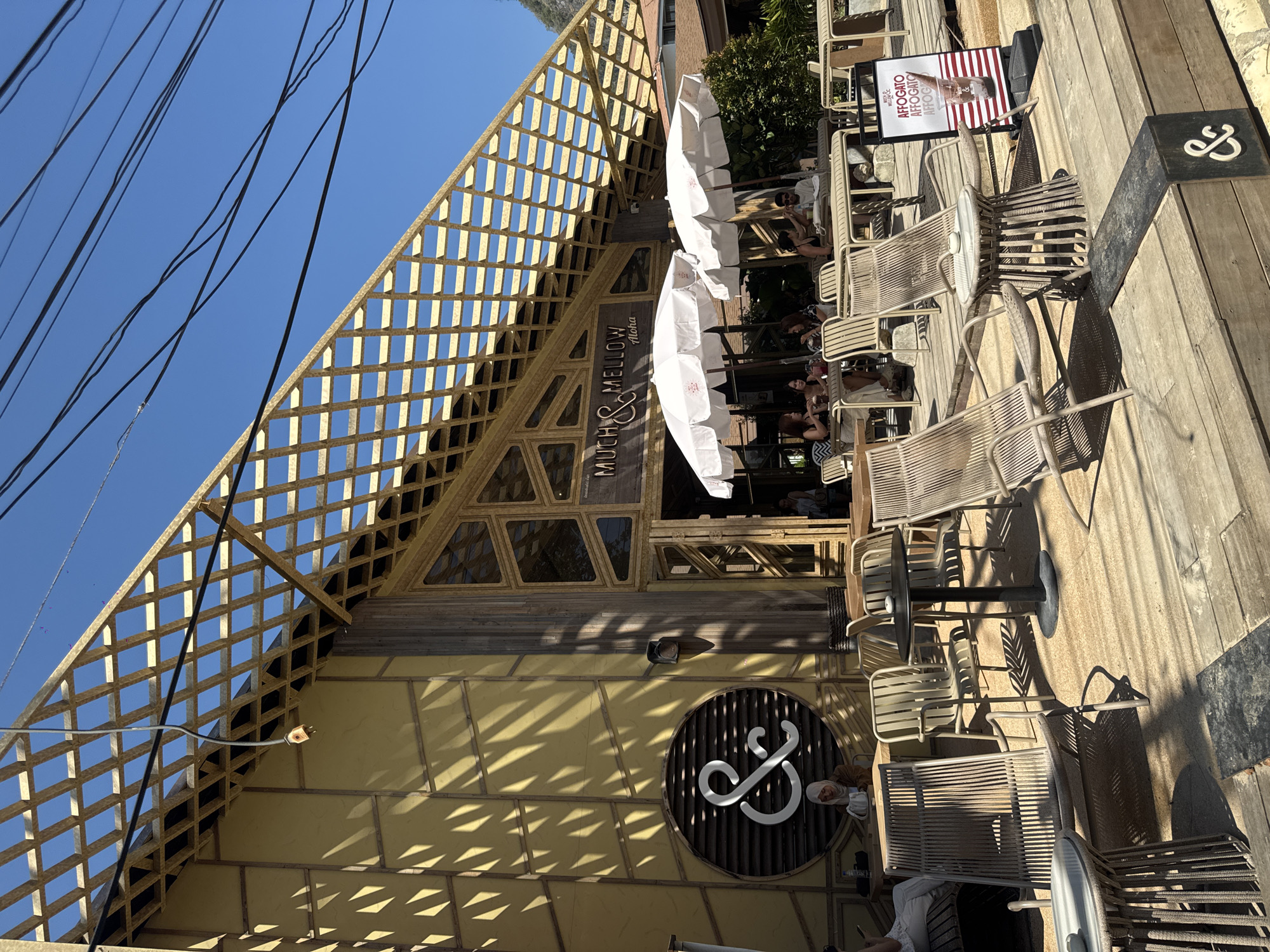 The exterior of "Much & Mellow" in Ban Ao Nang, Thailand is pictured during daylight. A triangular, latticed structure extends from the roof. Underneath, the building has a wooden facade, with a circular logo. Outside seating is arranged on a wooden deck, occupied by people under white umbrellas. Chairs and tables are visible. Power lines run above the structure. A sign advertising "AFFOGATO" is also visible.