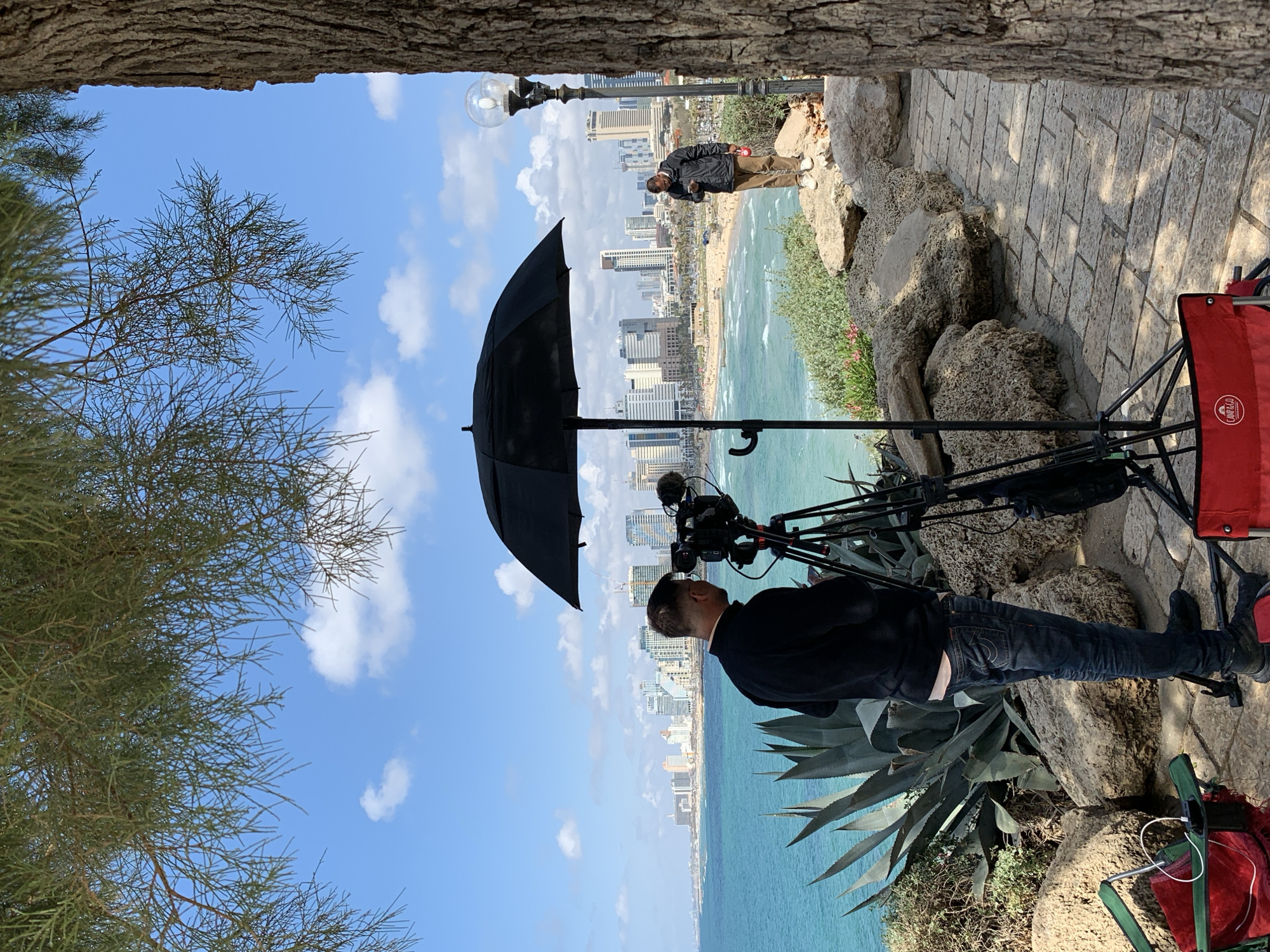 A photographer with a camera on a tripod, sheltered by a large black umbrella, is capturing a shot of the Tel Aviv coastline and skyline. The beach meets the turquoise sea, and buildings line the shore. Another person stands farther down the path, also observing the view. The scene is framed by trees and a stone wall, indicating a slightly elevated viewpoint, likely a park or observation point. A red folding chair and other equipment are visible near the photographer.