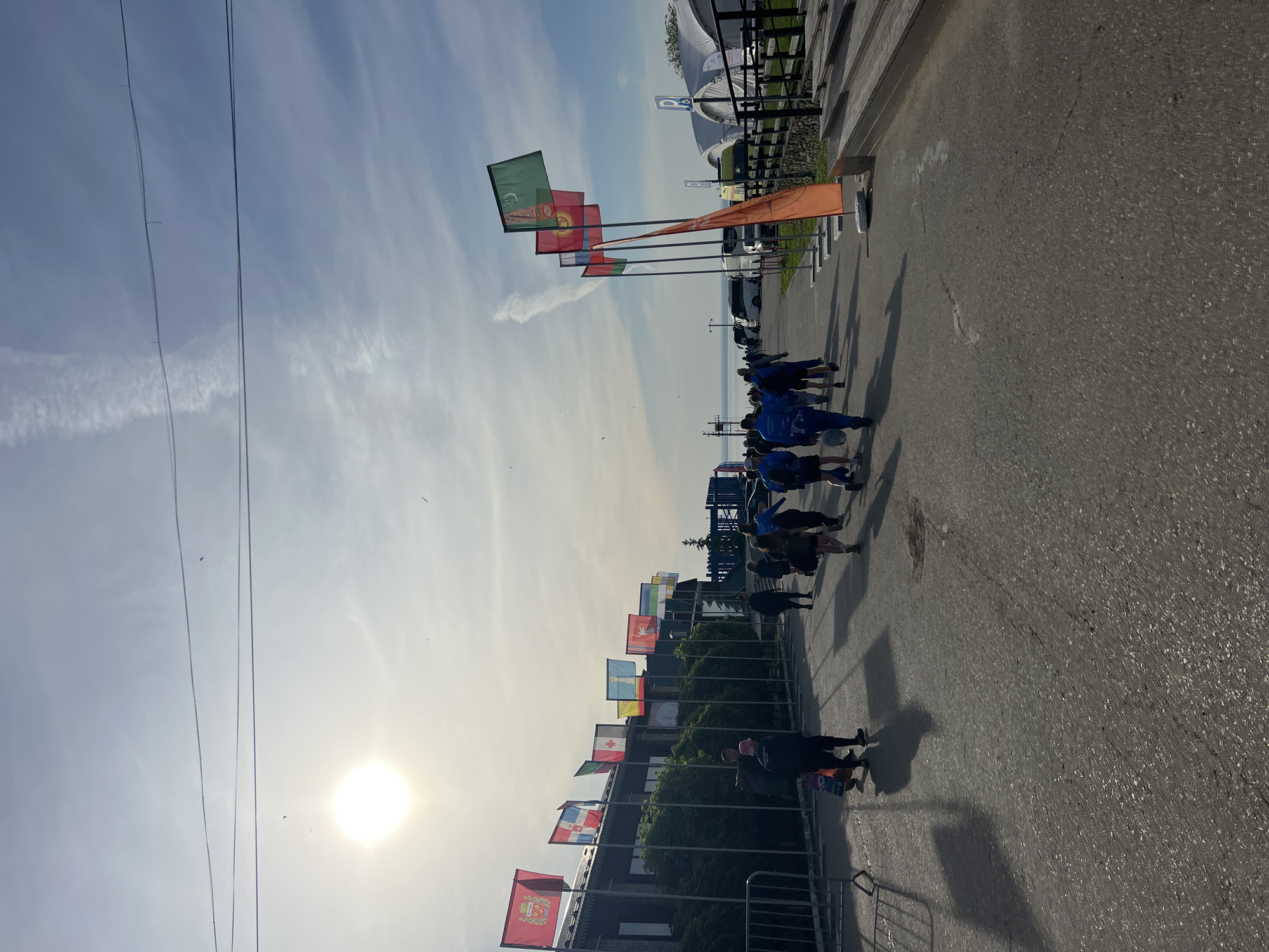 A vertical shot depicts a line of people walking along a paved road bordered by flagpoles. Flags of various nations are prominently displayed on both sides of the road. The setting sun is visible in the lower left corner of the frame, casting long shadows of the people and flags onto the pavement. The flags on the right of the image, closest to the camera, appear to represent nations such as France, Canada, and Russia. The flags on the left feature predominantly green and orange hues. A temporary metal barricade is visible in the lower left corner. A fence runs along the top right corner. In the background, tents and vehicles can be seen. The location is Khvalynsk, Russia.