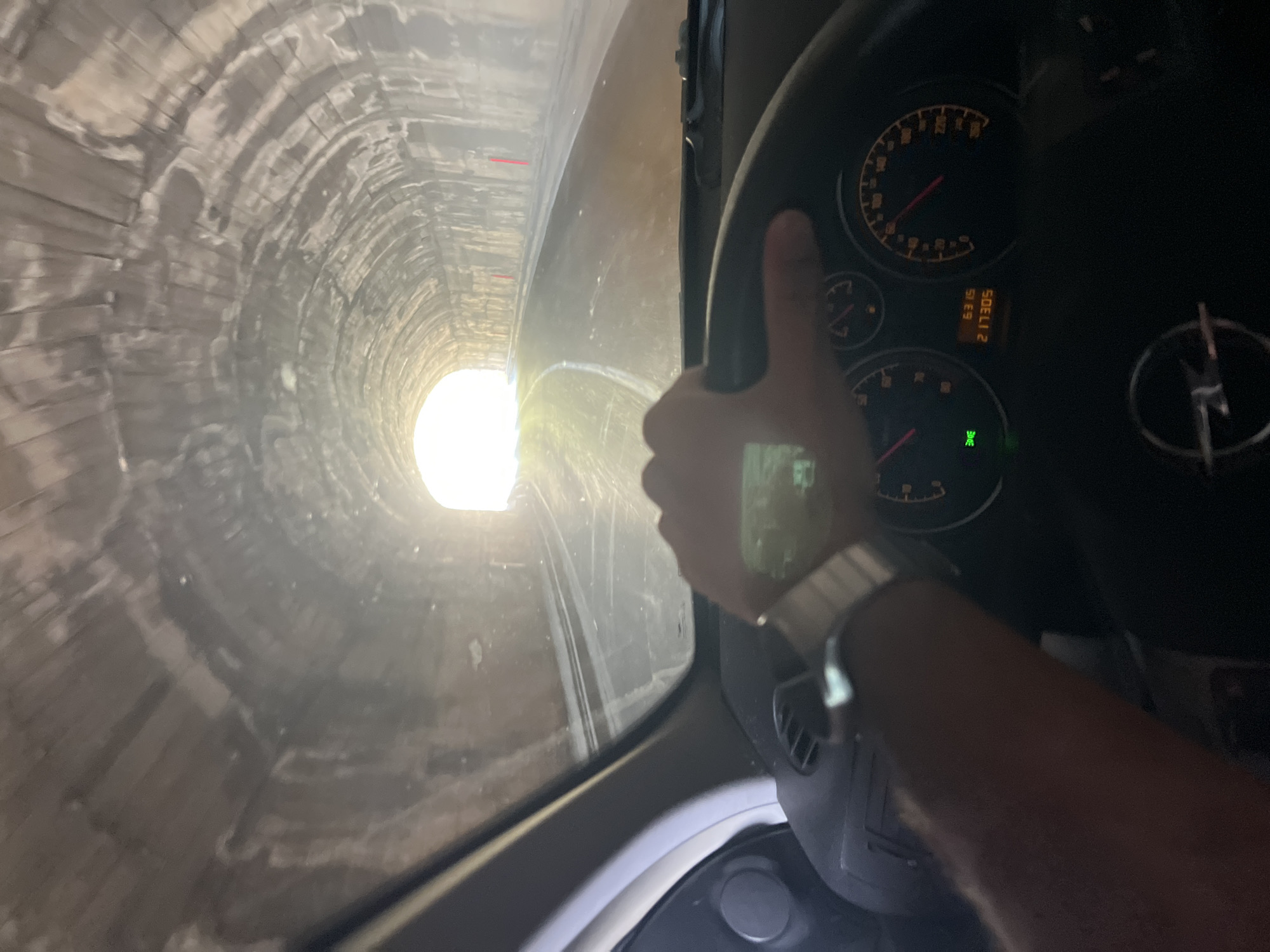 The image shows a first-person perspective of driving through a tunnel. The tunnel appears to be made of rough-hewn rock and concrete, with arched ceilings. The road ahead is dimly lit by the bright opening of the tunnel in the distance. The car's dashboard is visible, with a steering wheel, speedometer, and odometer displaying "6315" and "217305". A person's hand, wearing a watch, is on the steering wheel. The overall scene suggests movement and transit through an enclosed, man-made passage. This scene is consistent with the tunnel on the coastal road near Dobra Voda, Montenegro, which connects to the Adriatic Highway.