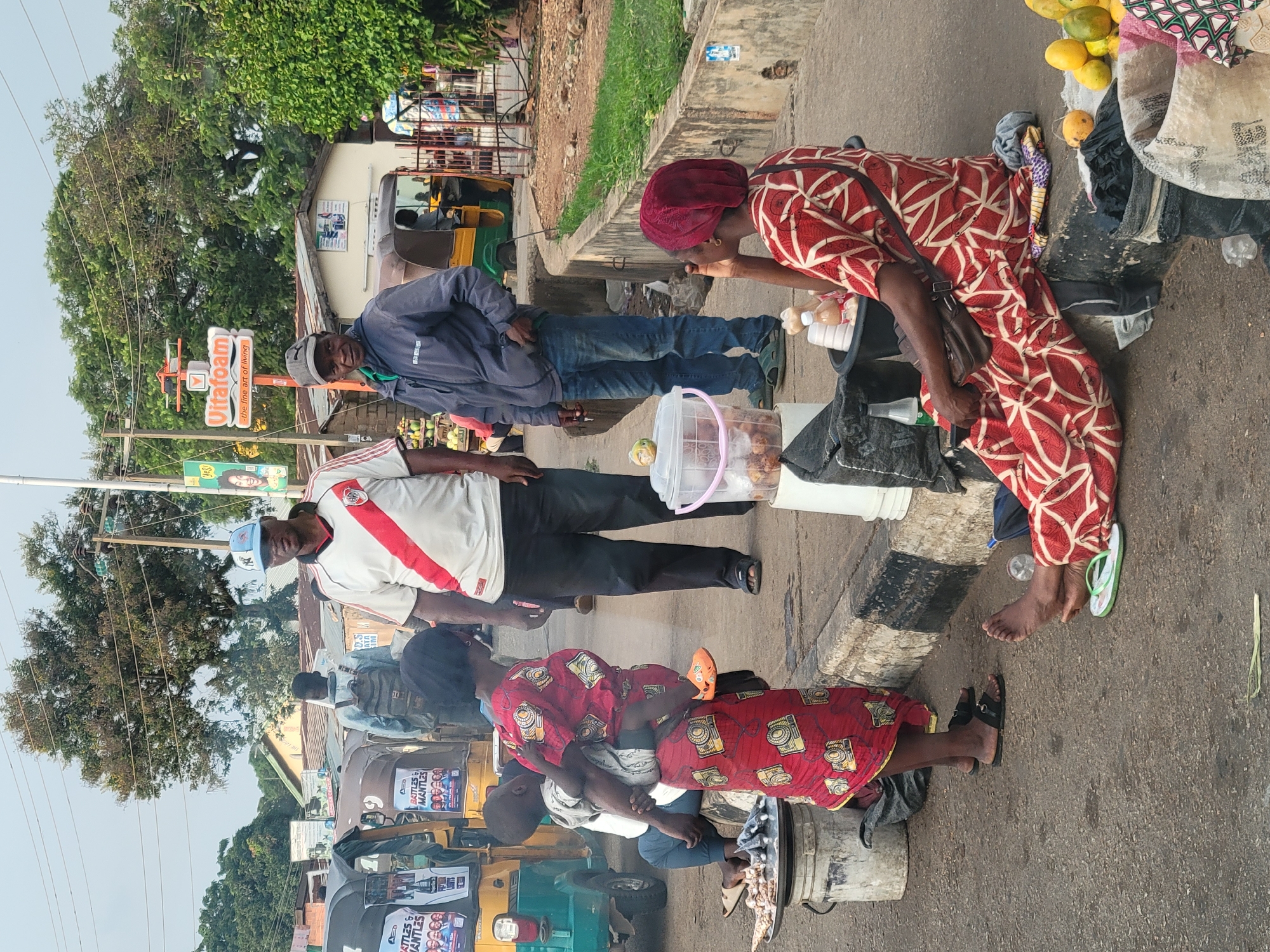 This is a street scene in Barakin Lerawi, Nigeria. A group of people are gathered on the side of a paved road. In the foreground, a woman wearing a red and white patterned dress and a red headscarf is sitting on a concrete curb, facing away from the camera, with her hand near her mouth. Beside her, a black bucket contains items, and in front of her are various fruits, including oranges and yellow fruits, and some bags. To her left, a woman in a red dress with a camera pattern is standing, carrying a child on her back. She is wearing black sandals. Next to her, a silver platter of food is placed on top of a white bucket. To the left of the platter is a green and yellow tuk-tuk with posters on its side.
Behind the standing woman, a man in a white and red striped shirt, black pants, and a blue and white baseball cap is facing the camera. He is standing next to another man wearing a grey hooded jacket and blue jeans, who is also facing the camera. Behind them, another man with a backpack is walking away. In the background, there are buildings, trees, power lines, and street signs, including a prominent sign for "Vitafoam - the fine art of living". Several other tuk-tuks are visible further down the street. The overall scene appears to be a busy marketplace or roadside vending area.