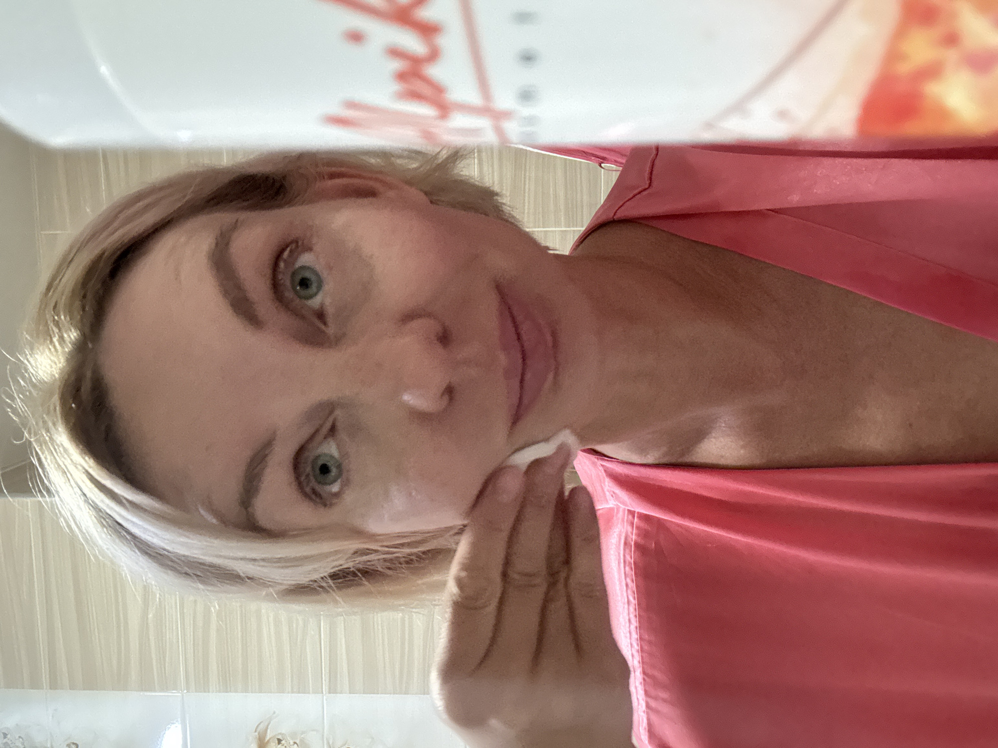 A close-up, selfie-style photograph shows a woman with blonde hair and blue eyes applying a product to her face with a cotton pad. She is wearing a coral-colored robe. A blurred white bottle with red lettering is visible on the right side of the frame. The background appears to be a tiled bathroom wall. There is no indication that this scene is located in Sochi, Russia.