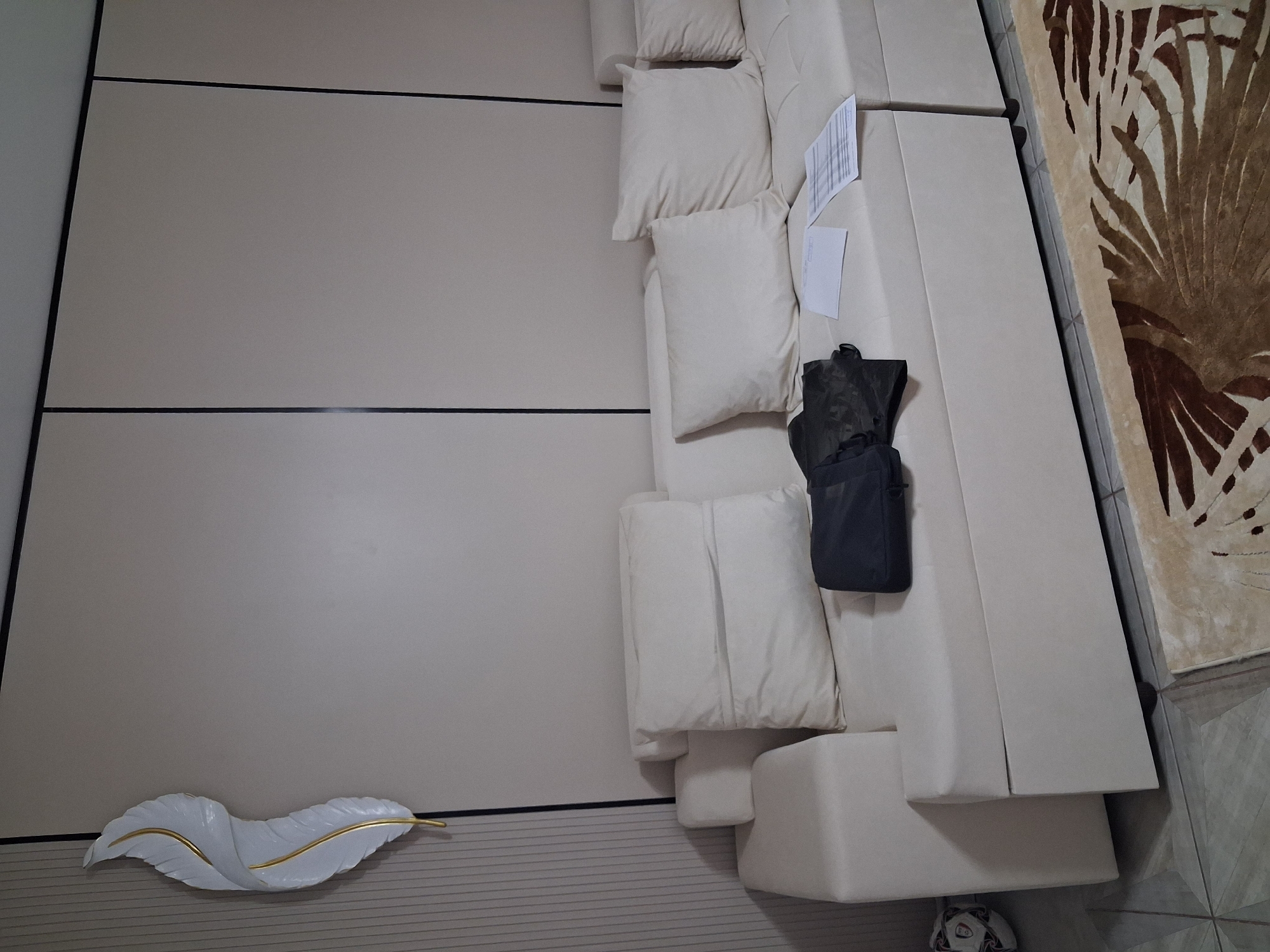 A white sectional sofa is positioned against a beige wall in what appears to be a living room. On the sofa, a dark bag and some papers are placed on the cushions. To the left of the sofa, a white and gold decorative feather-shaped wall art is mounted on a ribbed beige wall panel. In the lower left corner of the image, a soccer ball is visible on the floor. A patterned rug is partially visible in the foreground. The image is an indoor shot, likely from a residential setting in Guldala, Kazakhstan.