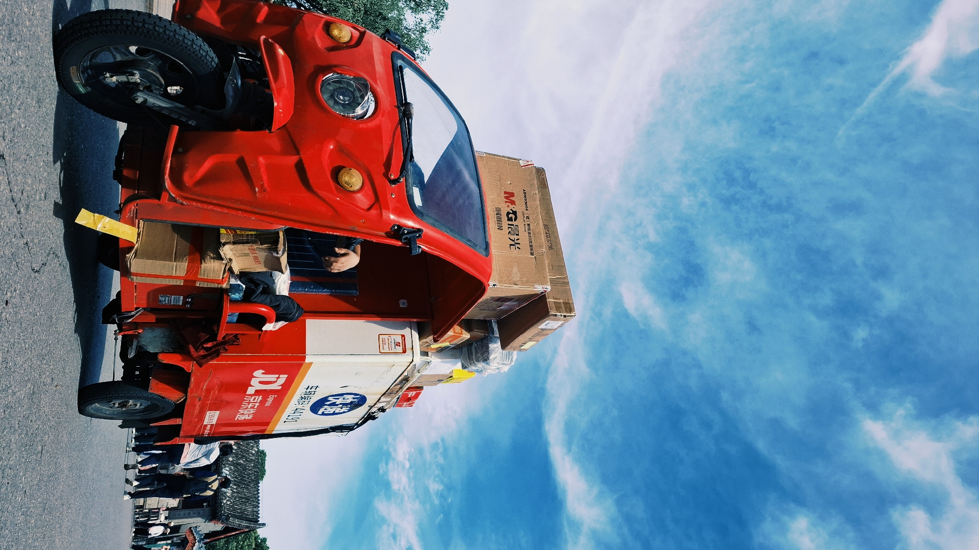 The image shows a red three-wheeled delivery vehicle parked on an asphalt road. The vehicle is heavily loaded with boxes, including one with the "MG晨光" (MG Chenguang) logo and the text "DISPLAY NEWRAN ADM95099(號)新悦派80页资料册 88 49.5 16." Another box has the "JD Express" logo and the text "车现编码:44191." The vehicle is partially visible, including the front wheel, cabin, and some of the cargo area. In the background, a group of people can be seen standing near a traditional Chinese-style building. The sky above is bright blue with wispy white clouds. The image is oriented vertically. Based on the presence of the JD Express delivery vehicle and the traditional architecture, the location is likely in Haidian District, China.
