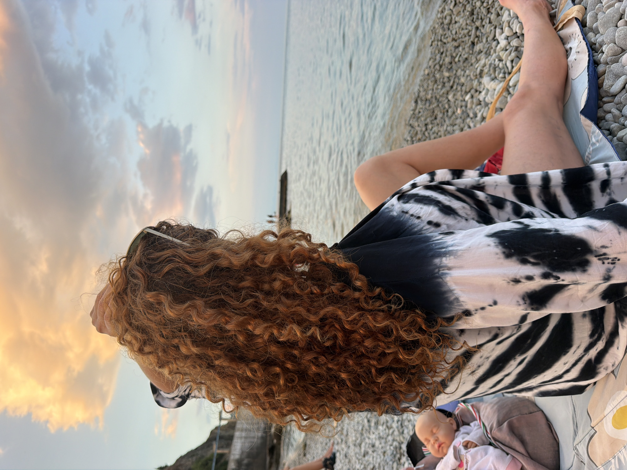 A woman with long, curly red hair is sitting on a pebble beach, looking out at the sea. She is wearing a tie-dye shirt with black and white patterns. To her left, a doll is lying on a blanket. In the background, there is a calm body of water and a cloudy sky with the colors of sunset. The environment suggests a peaceful moment by the sea.