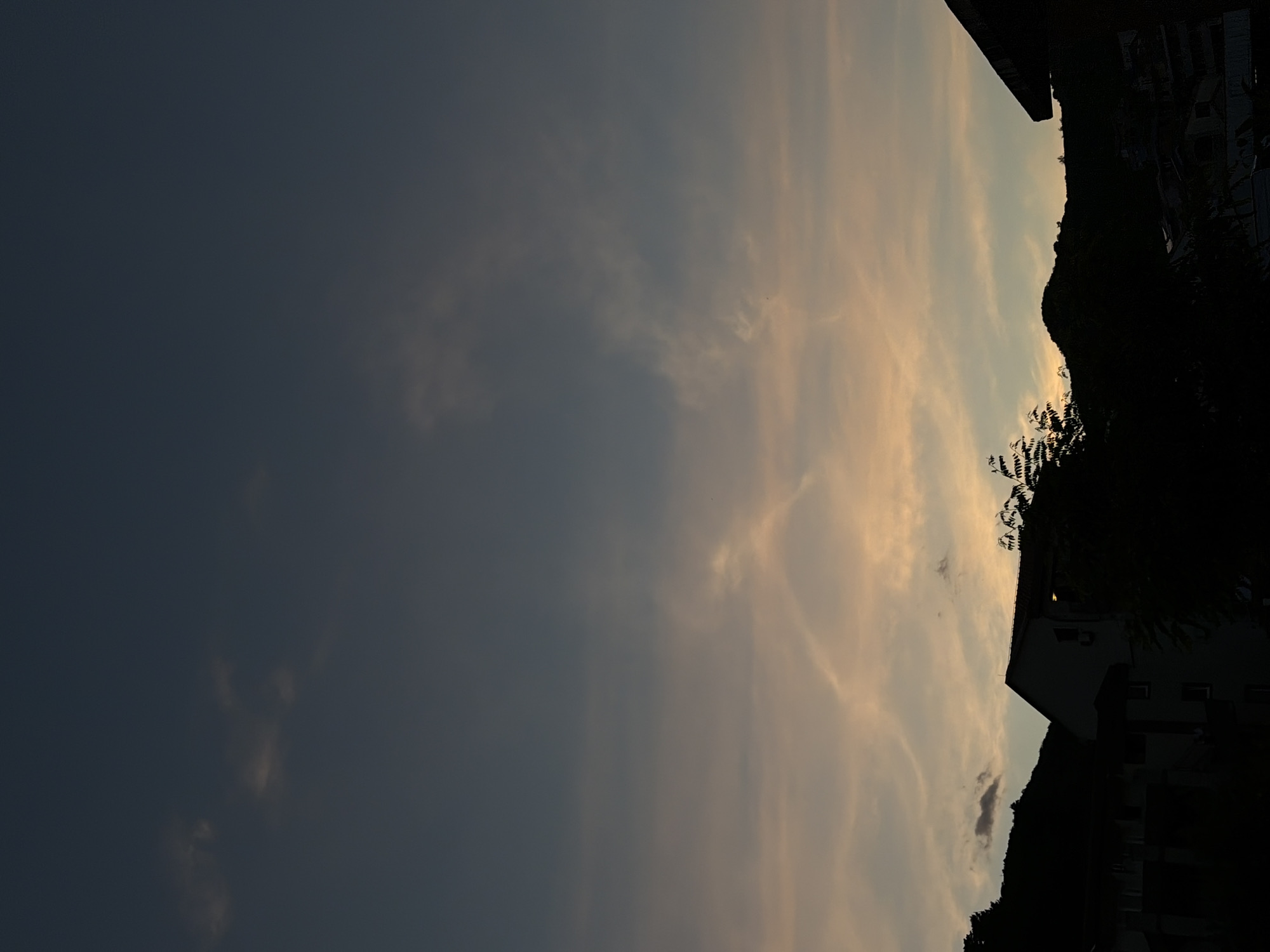 The image shows a dusk scene in Budva, Montenegro. The sky is a mix of dark blue and orange hues, with wispy clouds scattered throughout. Silhouetted against the sky are buildings and vegetation, including trees and foliage. The buildings are located on a hillside. The dark silhouette creates a contrast with the sky.