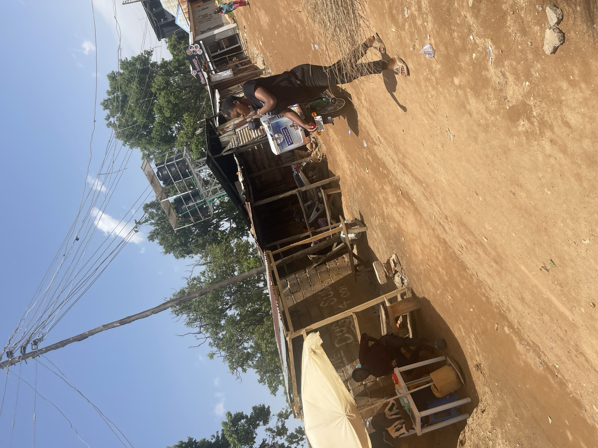 An outdoor daytime shot shows a street scene in Jos, Nigeria. The ground is dirt. A woman in black pants and a black t-shirt is walking away from the camera on the right, toward the background. She is wearing tan sandals. Behind her, to the left, a motorcycle is parked, with a white box on its back. A young woman is standing next to it, facing away from the camera. To the left of the motorcycle, a stall is set up with a beige umbrella, and two men are sitting at a table. The stall is made of wood and cinder blocks, with corrugated metal roofing. A power pole with many wires is visible in the background. The sky is blue with some white clouds. A building with signage that reads "Blessed Lounge" is visible in the background.