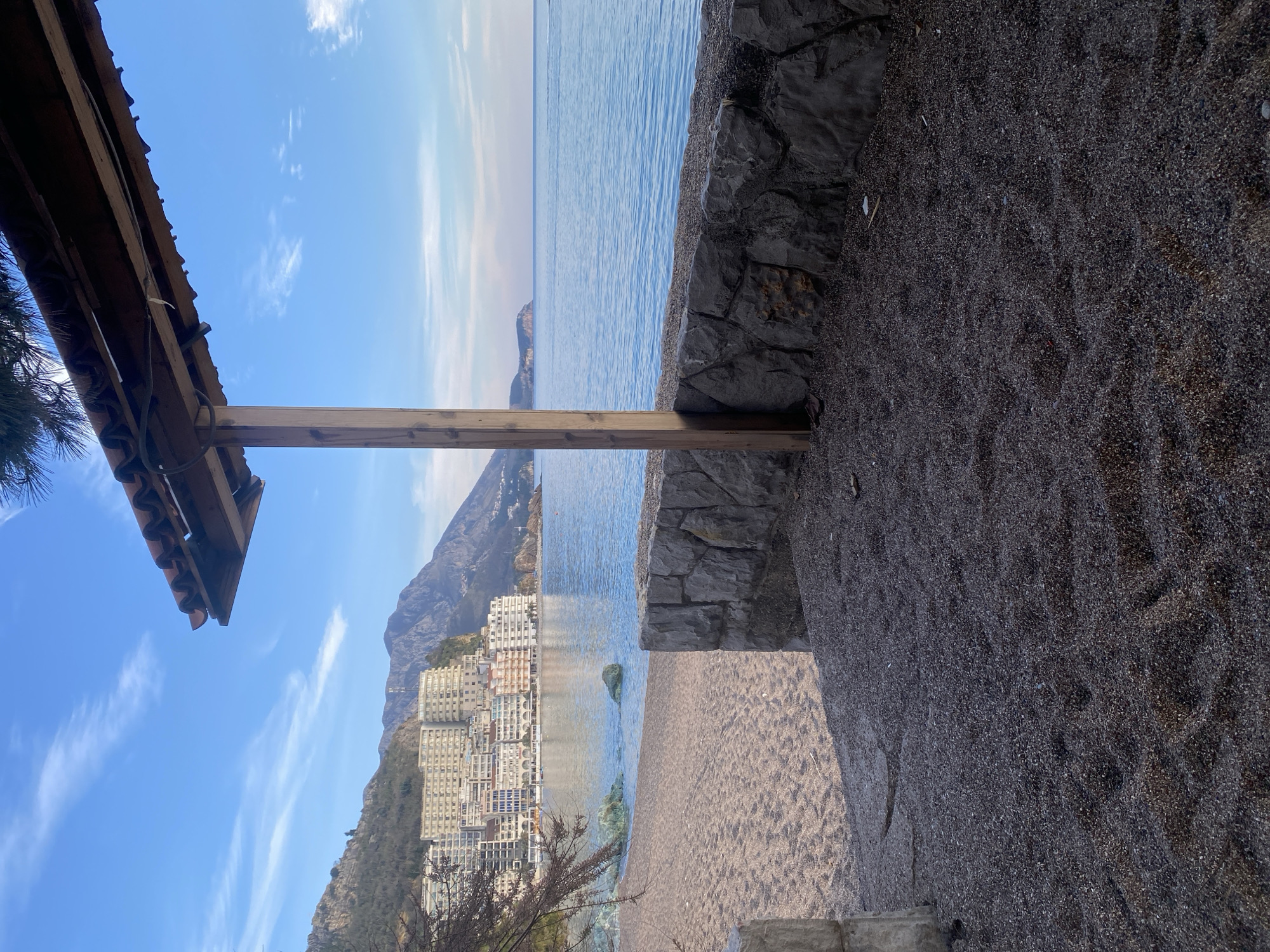 The image depicts a coastal scene in Rafailovići, Montenegro, from a vantage point under a partial shelter structure. The immediate foreground shows dark, coarse gravel with undulating patterns. To the right, a vertical wooden post rises, supporting an overhead structure with reddish-brown roof tiles, partially visible at the top right. A low, grey and brown irregular stone wall runs horizontally across the mid-foreground, separating the gravel from a light brown sandy beach.
The sandy beach curves along the coastline to the left, meeting the calm, light blue-green waters of the Adriatic Sea. Several light green rocks are visible in the shallow water near the shoreline on the left.
In the background, a densely packed cluster of multi-story buildings, predominantly white and light-colored, are terraced into the hillside on the left, characteristic of the Rafailovići townscape. Behind these buildings, rugged mountains, sparsely vegetated, extend across the horizon. The tranquil sea stretches to the far right, where a distant landmass or headland is visible. The sky is clear blue with scattered thin white wispy clouds. Bare tree branches are present on the far left, and pine needles are visible at the very top of the frame, suggesting overhead vegetation near the shelter. No human figures are present.