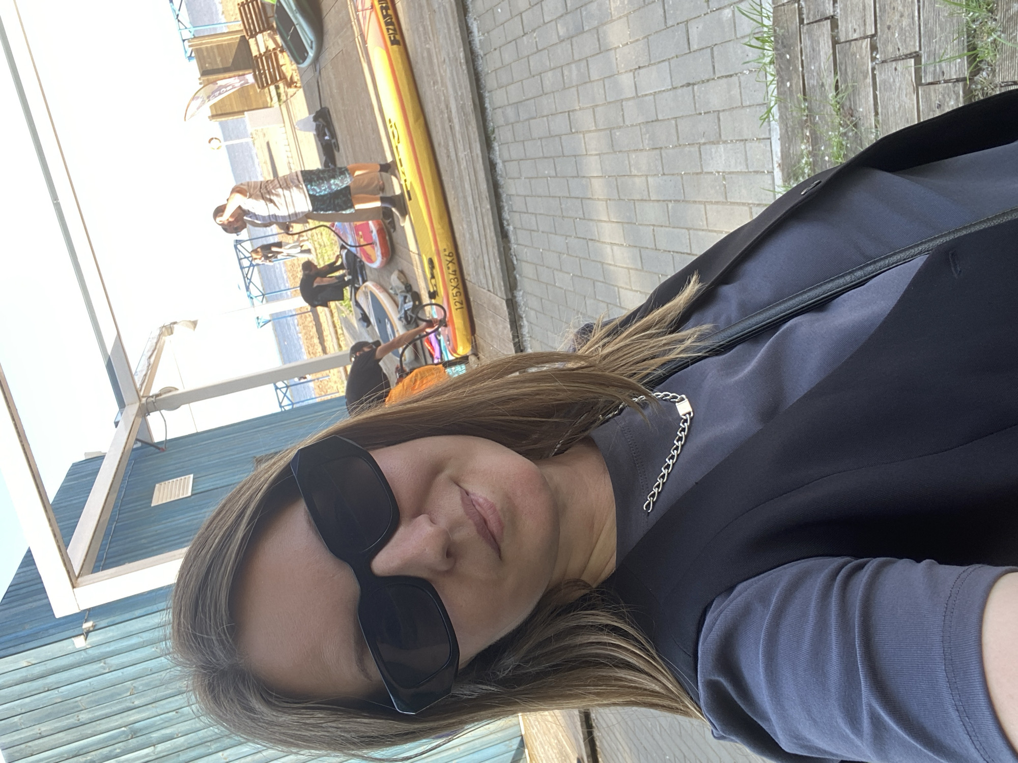 The image is a selfie taken by a woman wearing sunglasses, a dark vest, and a grey t-shirt with a silver chain necklace. She is standing outdoors, with a blue building on the left and a body of water in the background. Several people are visible in the background, some interacting with paddleboards. The ground is paved with bricks and wooden planks. The scene appears to be a waterfront recreational area, possibly in Chaikovsky, Russia, given the context.