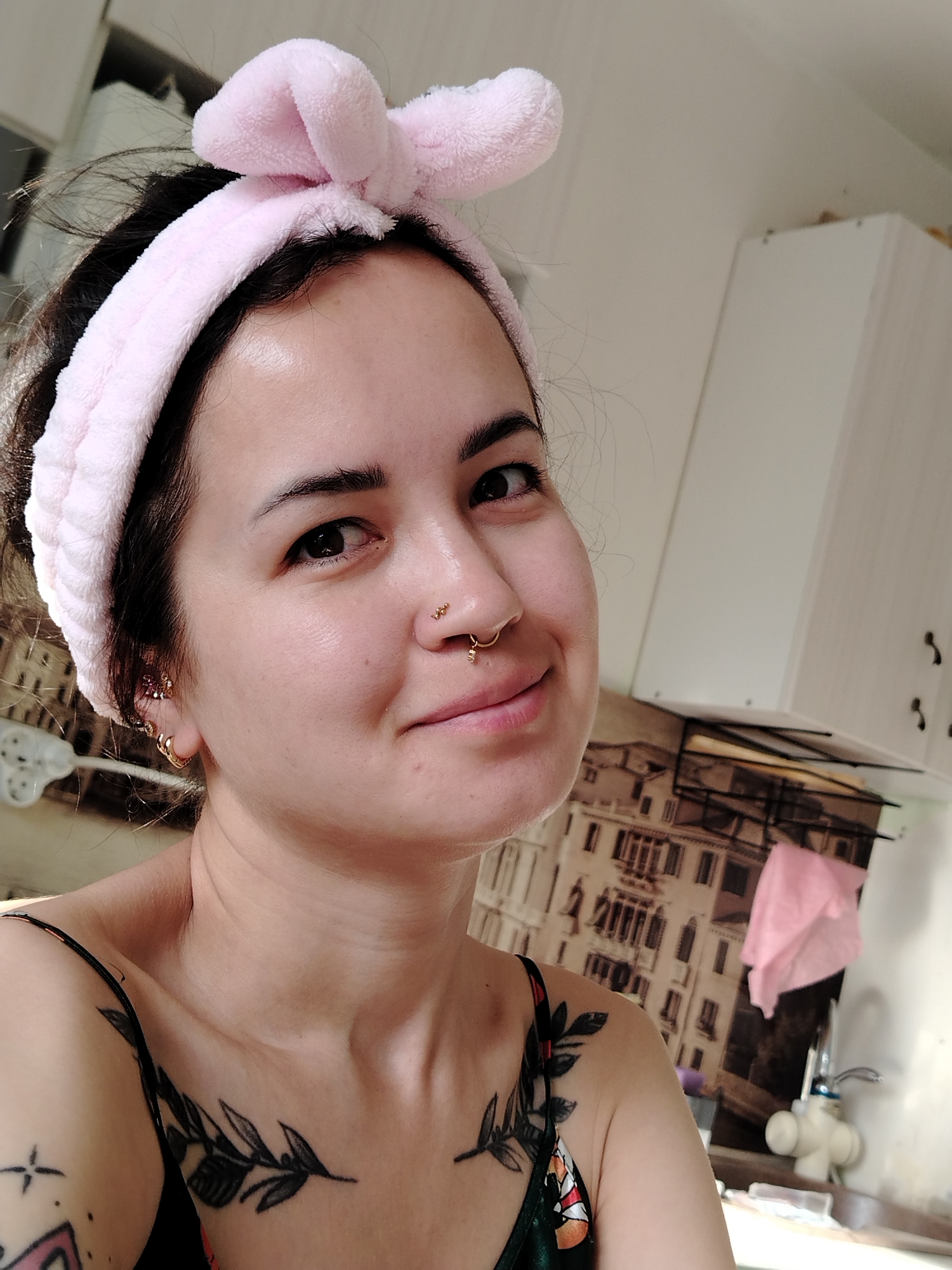 A young woman is depicted in a close-up, angled shot. She has dark hair pulled back by a light pink headband with bunny ears. Her skin is fair, with visible piercings in her nose and ear. She has a slight smile. She is wearing a tank top, exposing tattoos on her shoulder and chest. The background shows a kitchen setting with white cabinets and wallpaper depicting a cityscape. A faucet is visible in the background. No details indicate this scene is in the city of Noviy, Russia.