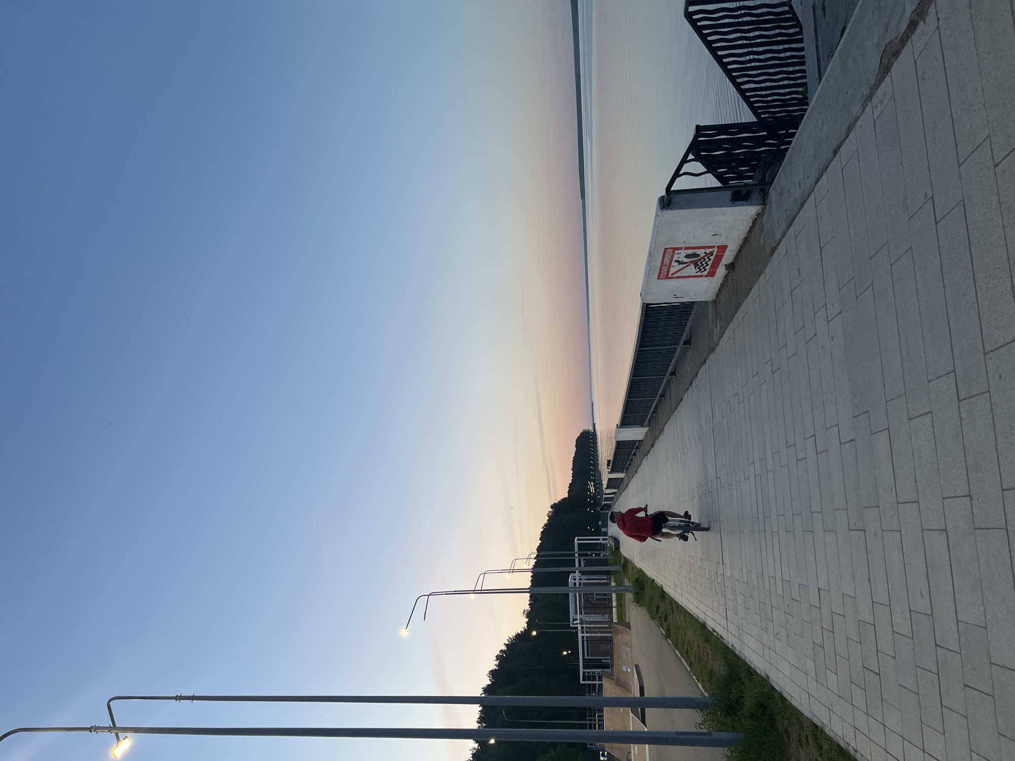 A cyclist in a red jacket is riding on a paved path alongside a body of water at sunset. The path is bordered by a metal railing and a road with streetlights. The sky is clear with a gradient of blue to orange near the horizon. The location is likely Chaikovsky, Russia.