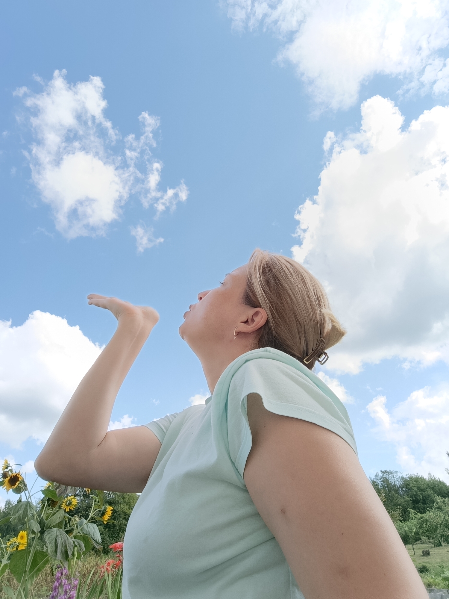 A woman with blonde hair pulled back, wearing a light blue t-shirt, is seen from the chest up, outdoors. She is facing upwards and to the left, with her mouth puckered as if blowing a kiss. Her left hand is extended towards the sky. In the background, there is a bright blue sky with scattered white clouds. Below the sky, a field of flowers, including sunflowers, is visible, along with green trees. The setting appears to be a rural or garden area. There is no indication of the city of Dubovaya, Russia within the image.
