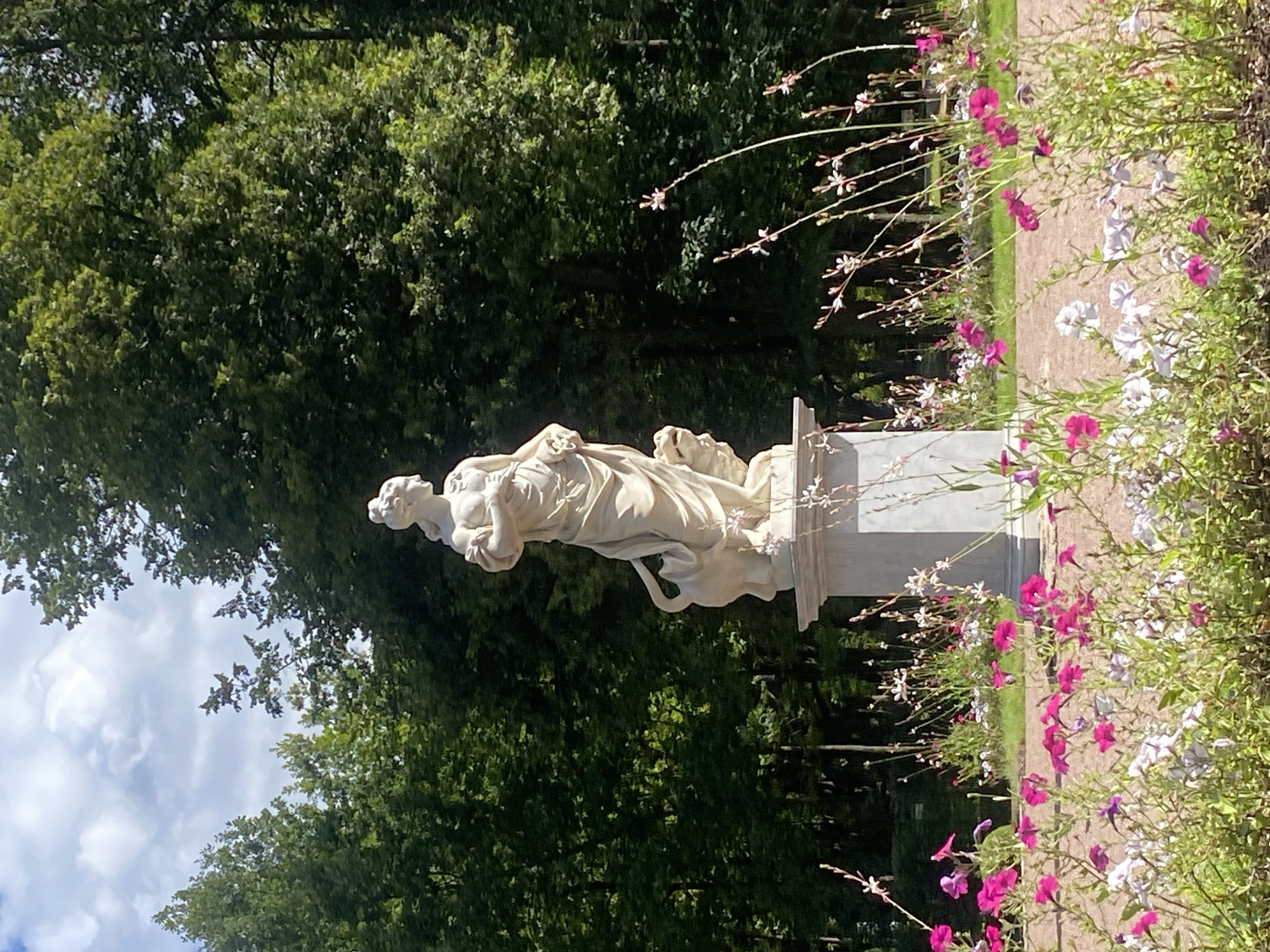 A pale stone statue of a female figure stands on a pedestal in a park, surrounded by a variety of flowers. The statue depicts a woman with flowing robes, holding a staff in her left hand and with a lion at her side. Behind the statue, a dense wall of green trees forms the backdrop. The foreground is filled with a vibrant display of pink and white flowers, with some thin, delicate pink flowers reaching upwards. The ground appears to be a gravel path. The sky above is a mix of blue and white clouds. This scene is located in the Pavlovsk Park, Pavlovsk, Russia.