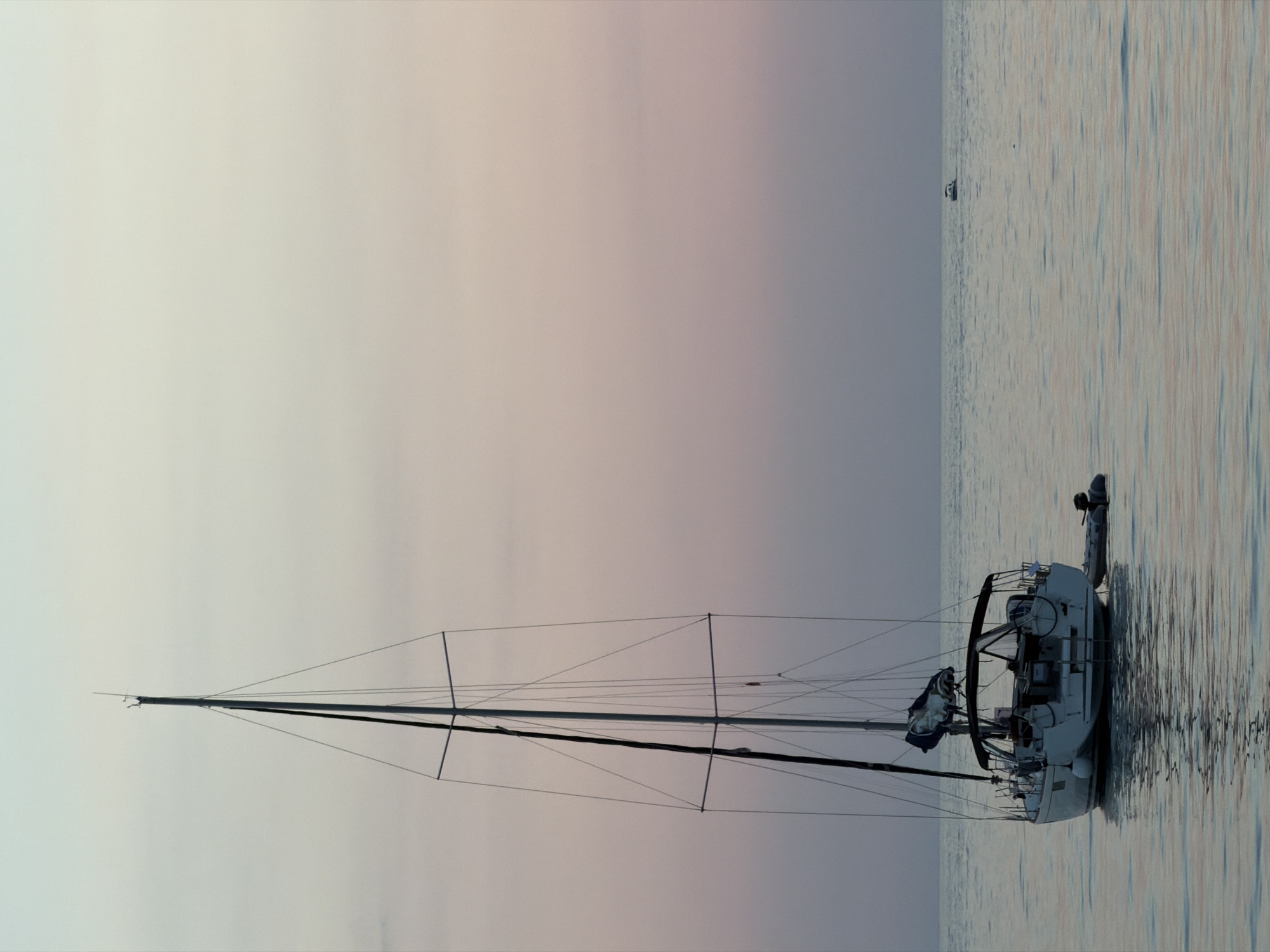 A single sailboat is anchored in the calm, shimmering waters of the Adriatic Sea at dusk, its mast tall and thin against a soft gradient of pale blue and pink sky. The boat is white with a dark canopy over the cockpit and a small inflatable dinghy attached to its stern. In the distance, on the horizon, a small motorboat is visible. The water reflects the muted colors of the sky and the silhouette of the sailboat. The scene suggests a peaceful evening at sea, likely off the coast of Bar, Montenegro, given the context.